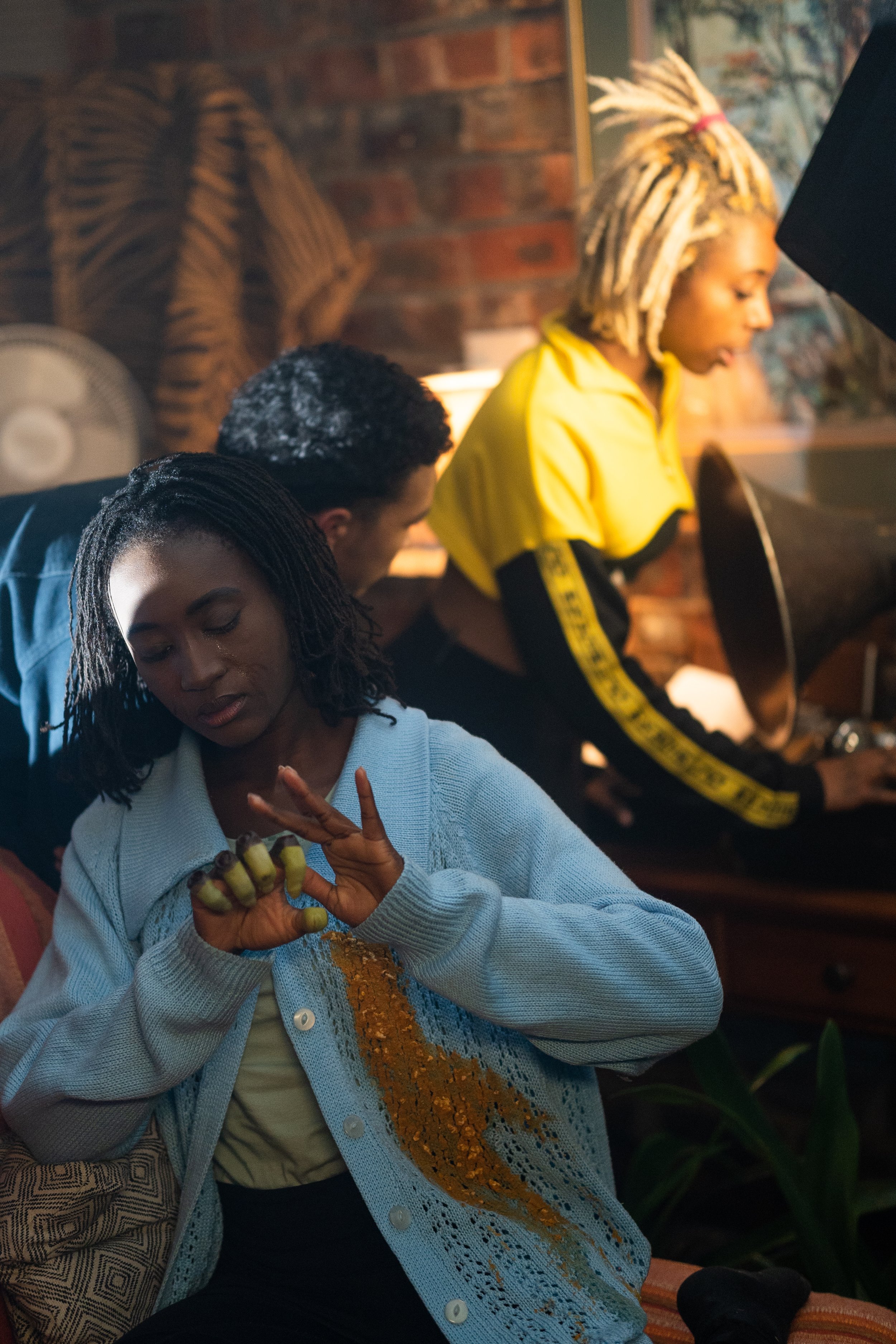 A woman with dreadlocks wearing a light blue sweater with yellow sequins, sitting and looking at her hand, while two other women with curly and dreadlocked hair work at a vintage typewriter in a cozy room with brick walls.
