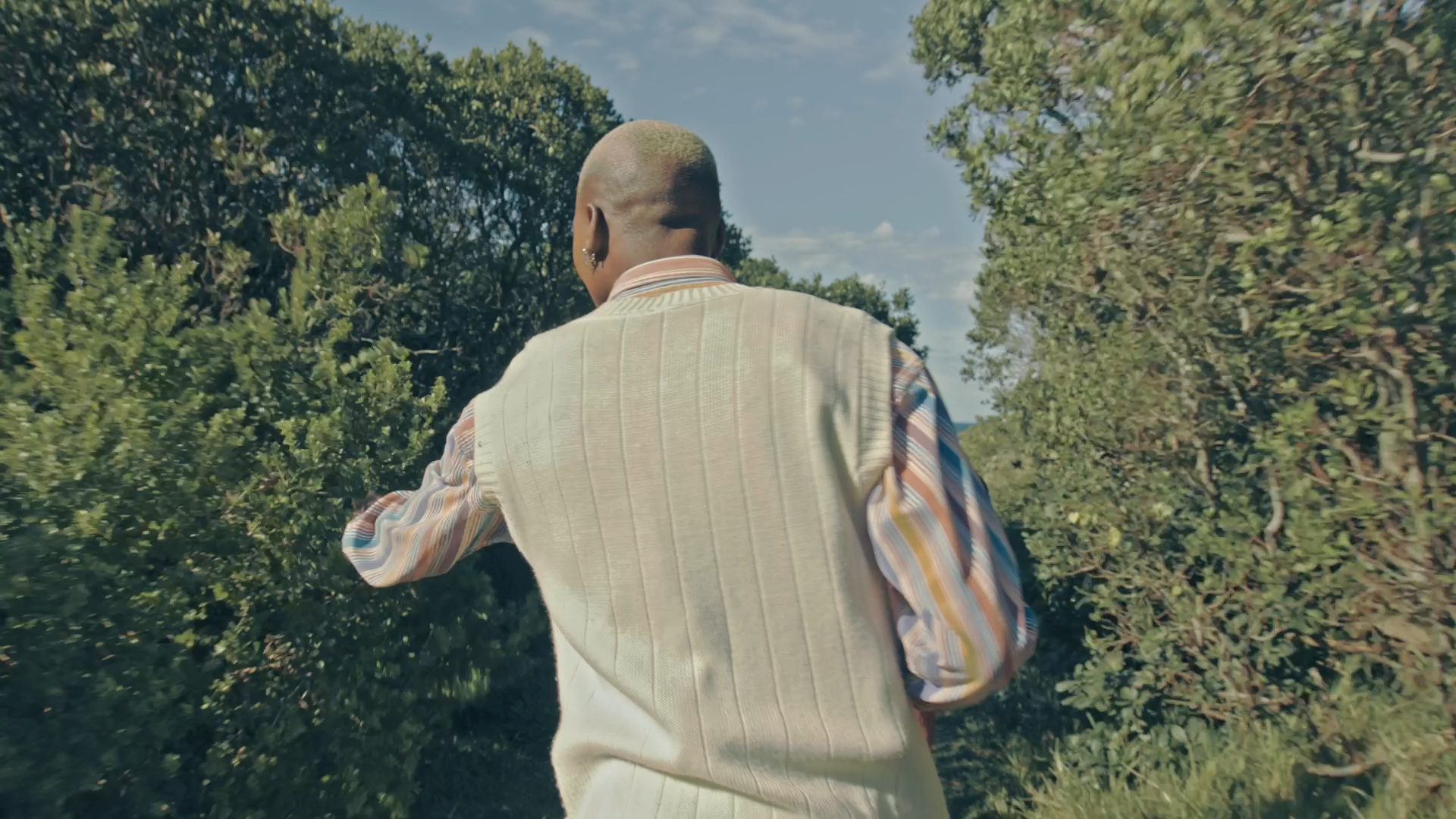 Person with short hair, earrings, and striped shirt under a light vest, walking through a lush, green outdoor area with trees and bushes on a sunny day.