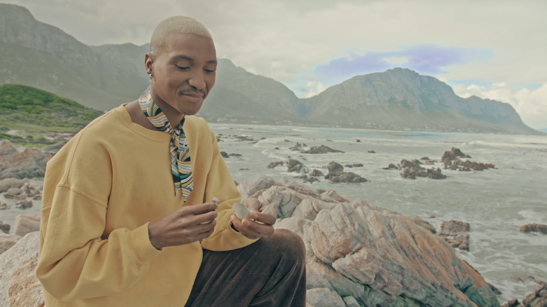 A person with a short blonde haircut, wearing a yellow sweatshirt and a patterned scarf, sitting on rocks by the ocean with mountains in the background, holding a small flower and a smartphone.