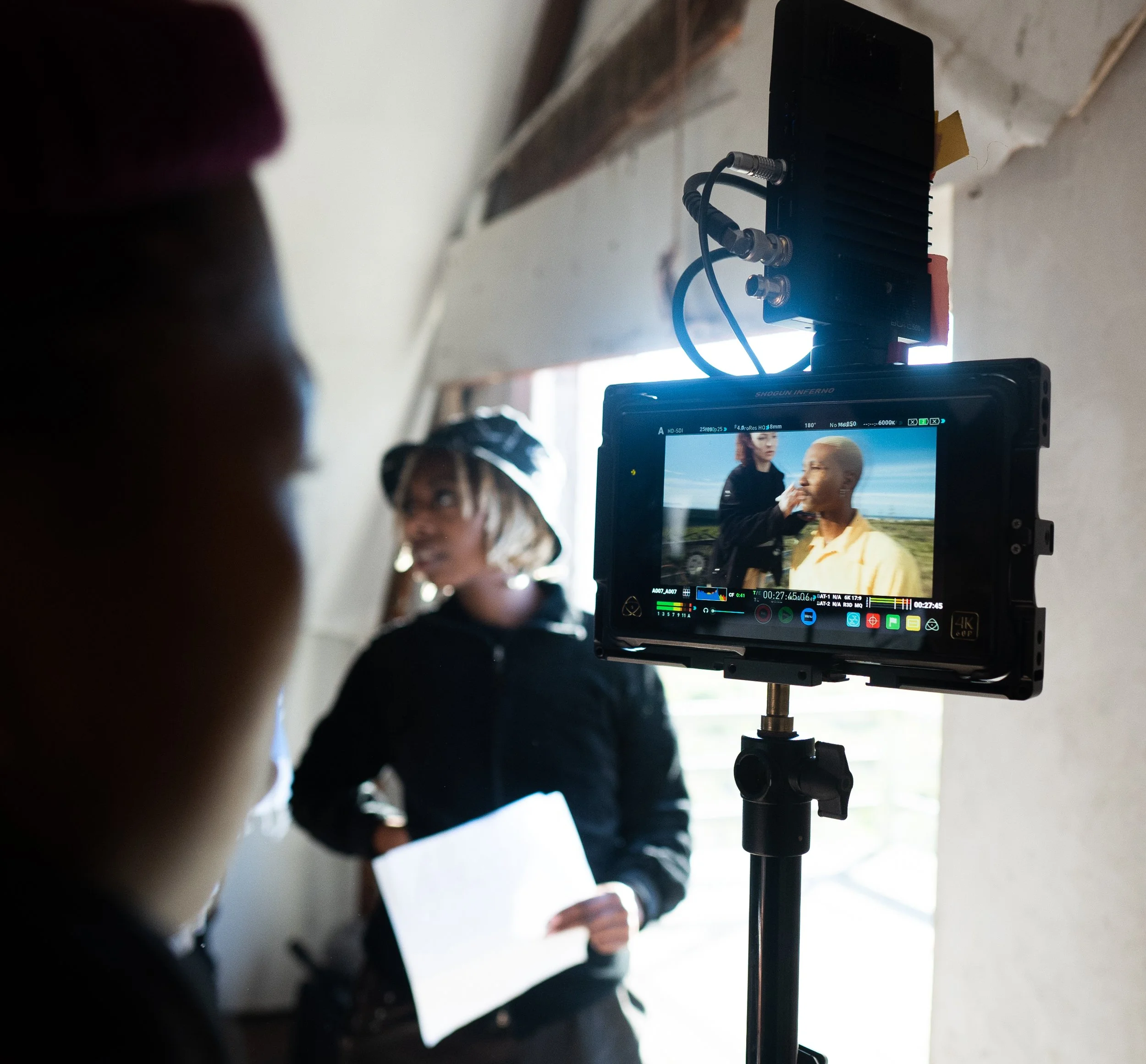 Behind the scenes of a film or video production, a monitor displays a shot of two women, one with short hair and a shaved head, talking outdoors. One woman is touching the other's face, and the person recording is holding a script or notes.