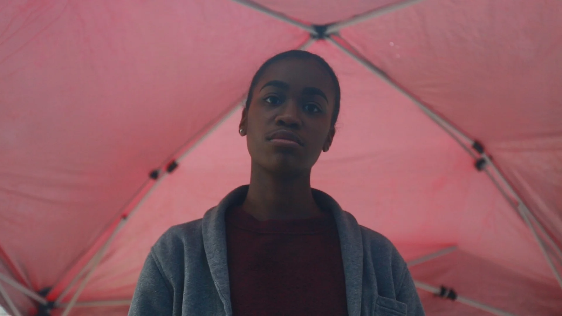 A young woman under a pink umbrella, looking at the camera with a serious expression.