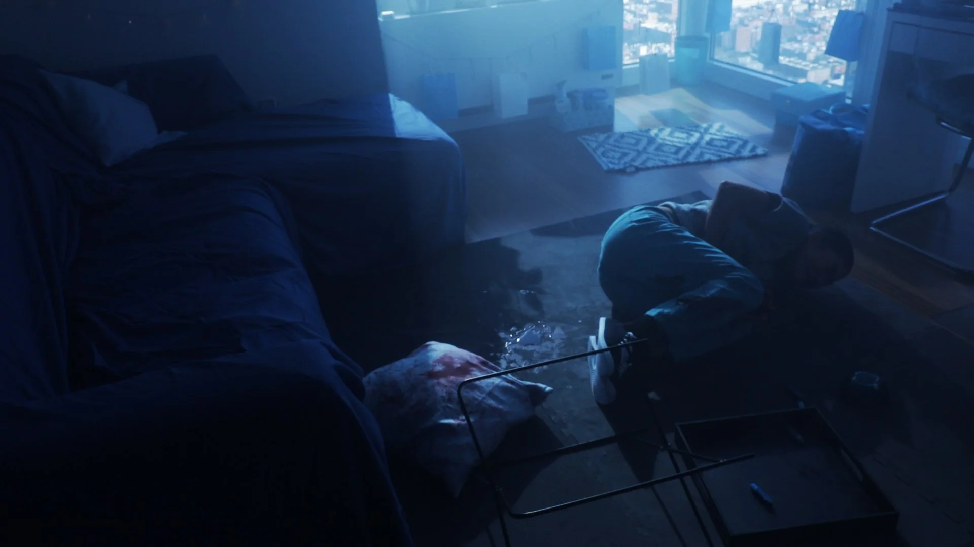 Person crouching on the floor in a dimly lit living room during daytime, with a glass coffee table and a blue couch nearby. The room has large windows showing a cityscape outside.