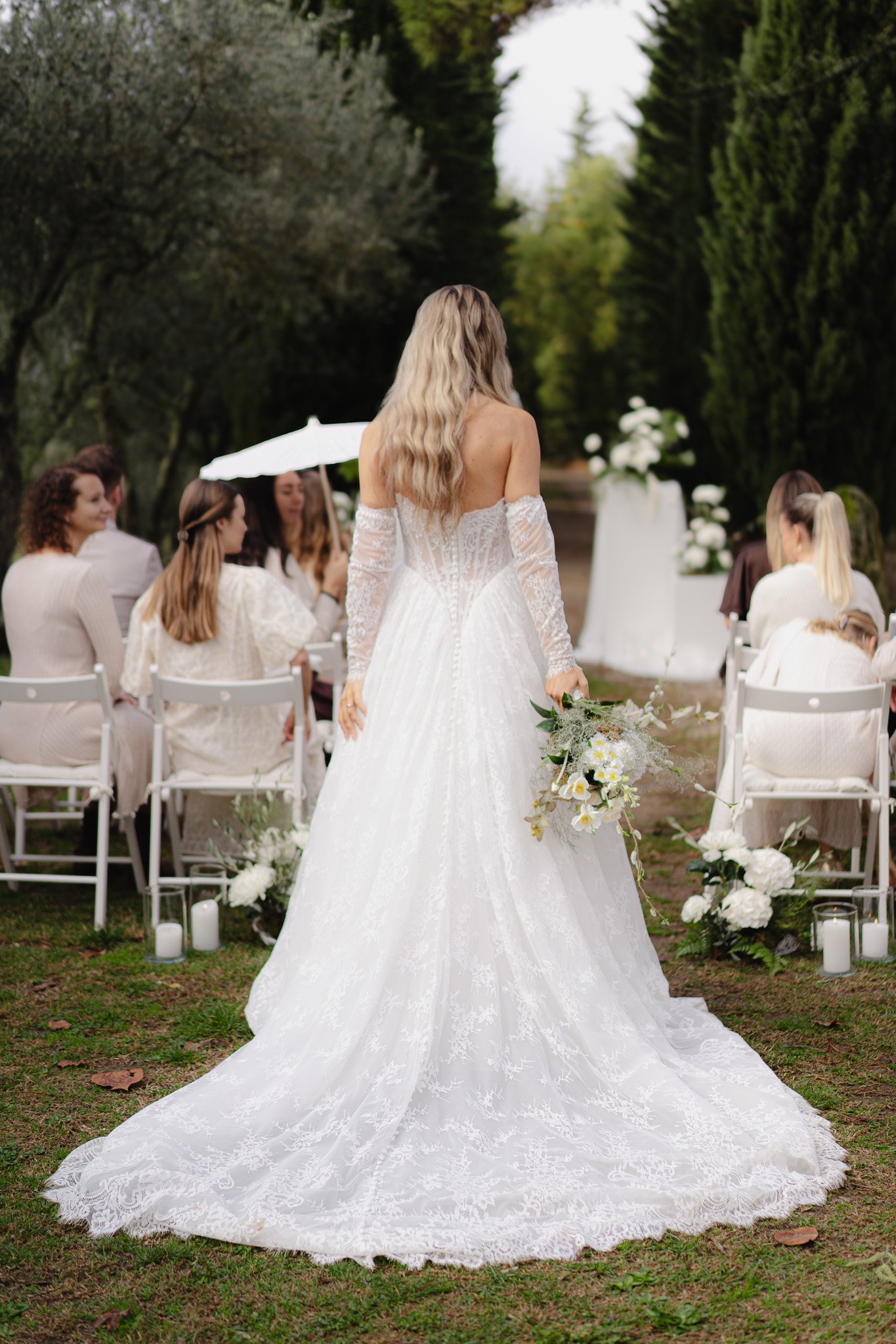 Braut in einem weißen, langem Hochzeitskleid mit Spitze, steht bei einer Hochzeitszeremonie im Freien, hält einen Blumenstrauß, umgeben von Gästen, die auf weißen Stühlen sitzen, und Dekoration mit weißen Blumen und Kerzen.