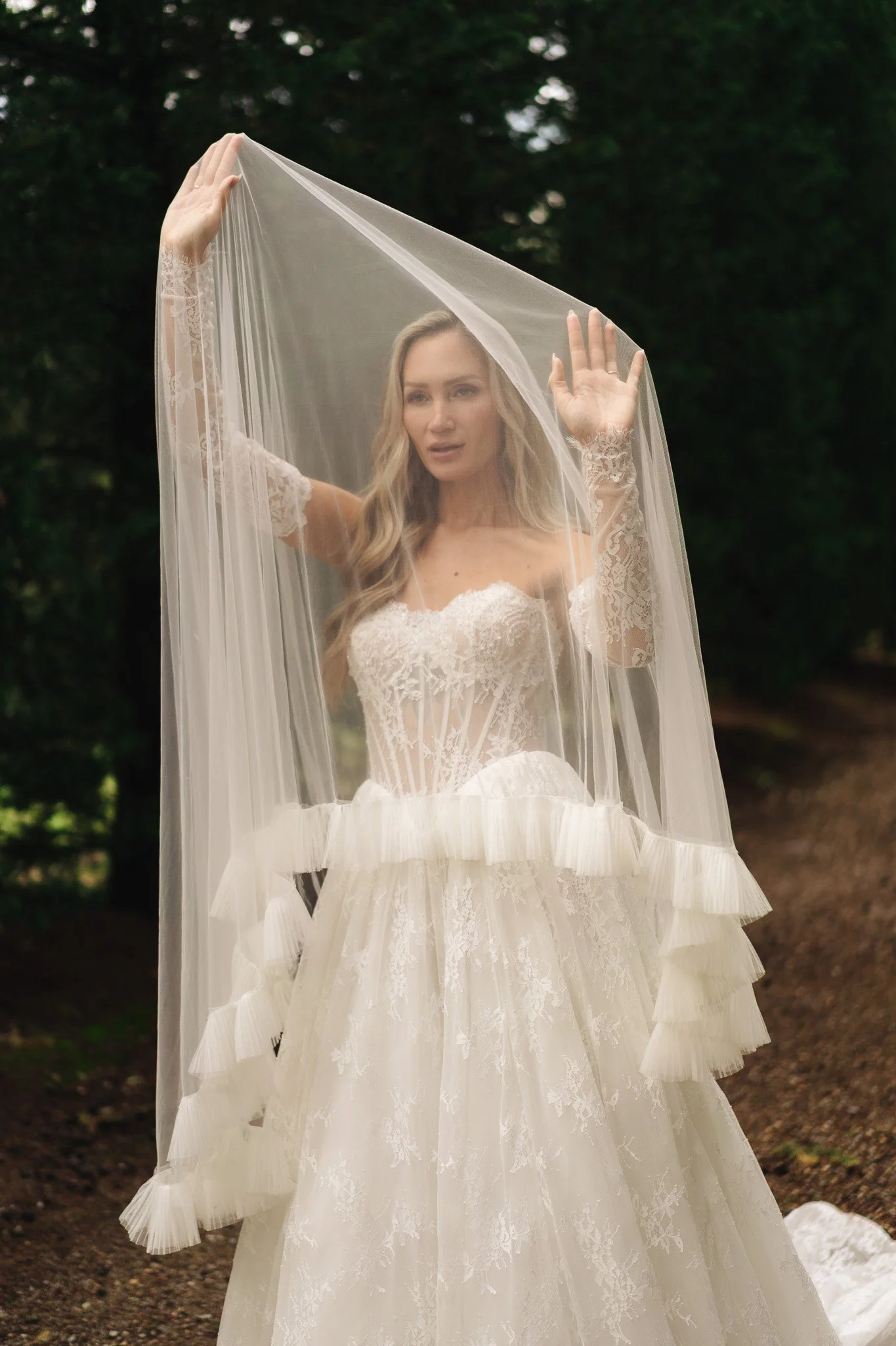 Eine Frau in einem weißen Brautkleid, die einen Tüllschleier hält, vor einem dunklen, bewaldeten Hintergrund.