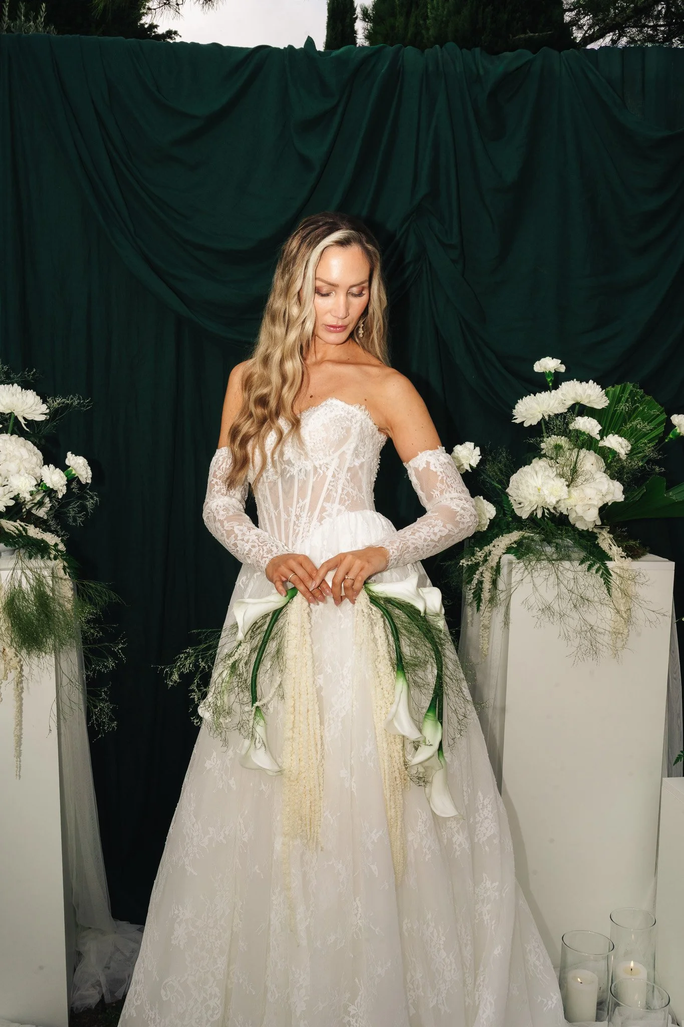 Eine Frau in einem weißen Hochzeitskleid mit Spitze steht vor einem grünen Hintergrund, umgeben von weißen Blumen und Kerzen. Es scheint eine Hochzeitsfeier zu sein.