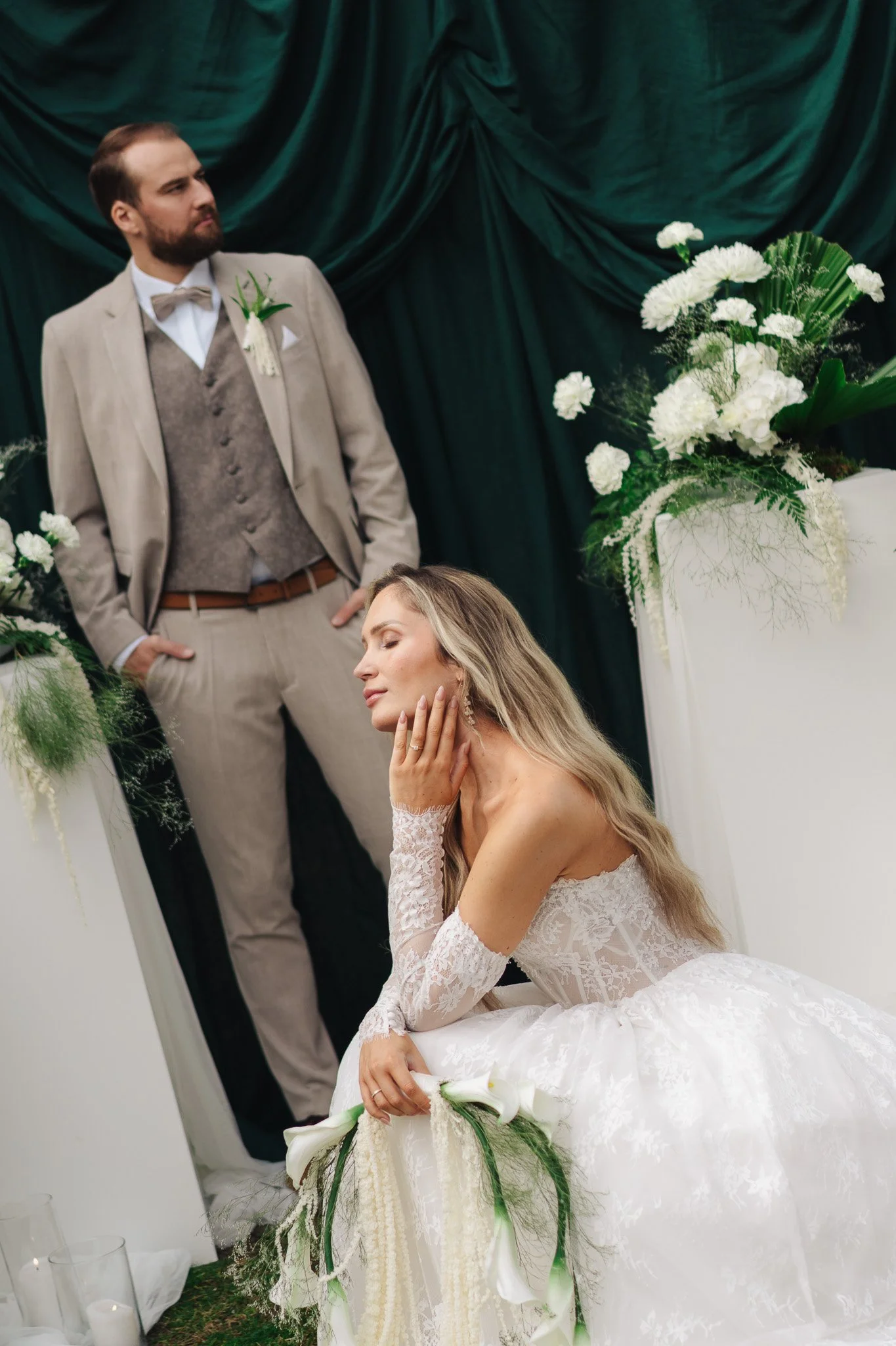 Ein Mann in Brautkleid mit einem Hochzeitsanzug, der vor schwarzem Satin hängt, sowie eine große weiße Blumenarrangement auf einem Podest.