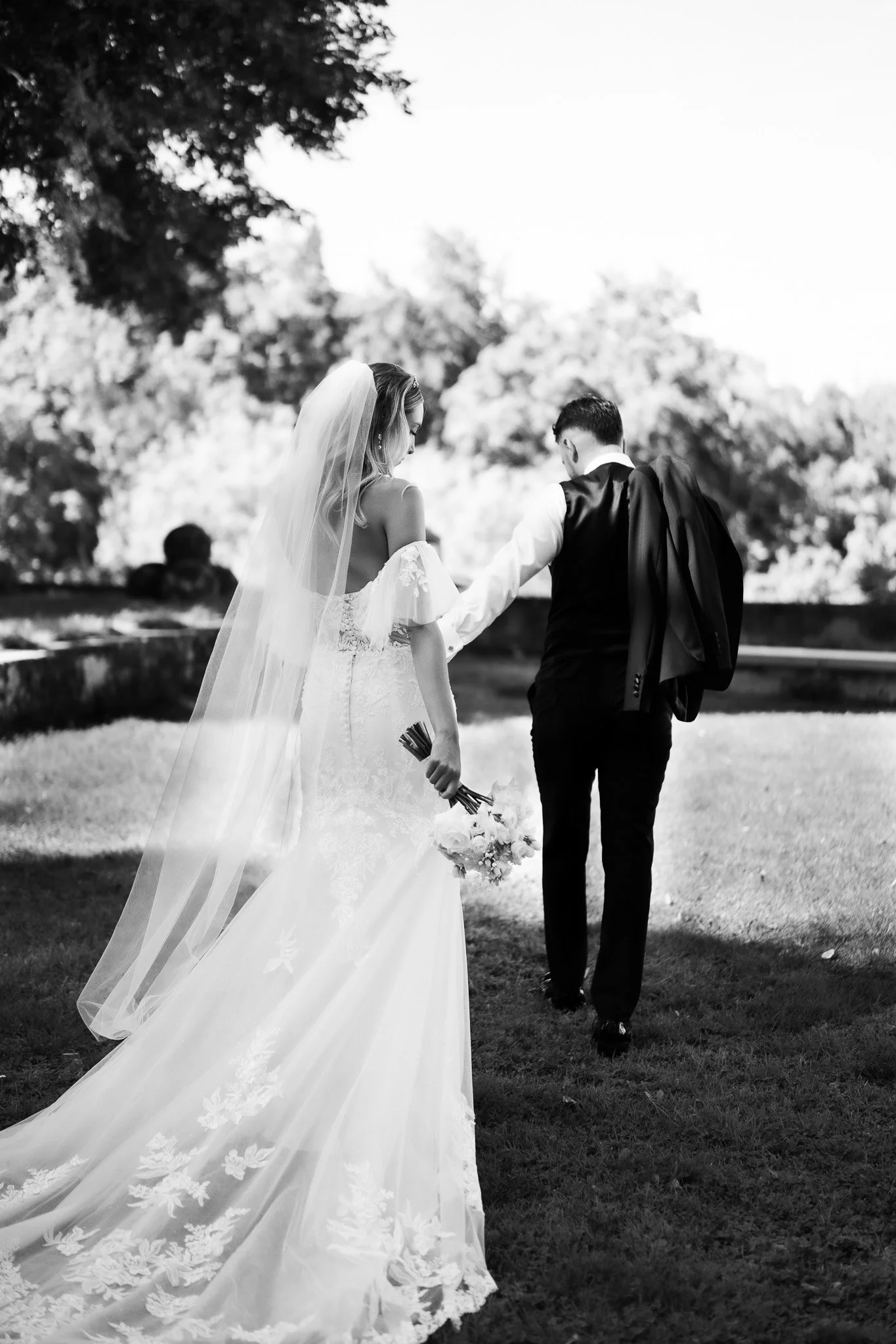 Ein Brautpaar beim Spazieren im Park, die Braut trägt ein weißes Hochzeitskleid mit Schleier und hält einen Blumenstrauß, der Bräutigam trägt Anzug mit Sakko, trägt es über der Schulter.