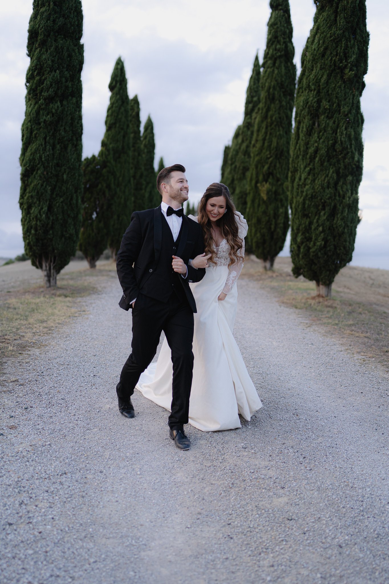 Ein frisch verheiratetes Paar spaziert auf einer Steinweg zwischen hohen, schmalen Tannenbäumen im Freien, beide lachen und tragen formelle Kleidung.