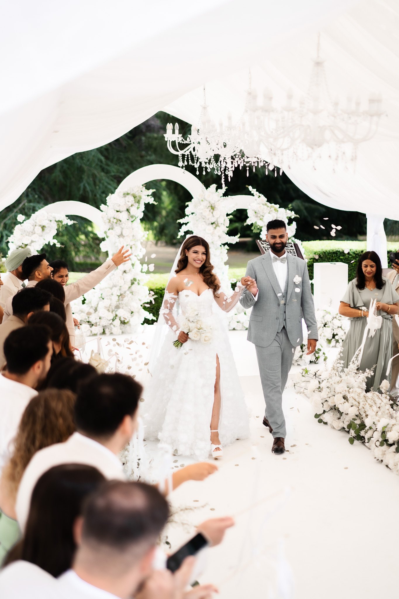 Ein Brautpaar bei einer Hochzeitszeremonie in einem weißen Festzelt, umgeben von weißen Blumen und Gästen, die applaudieren und feiern.