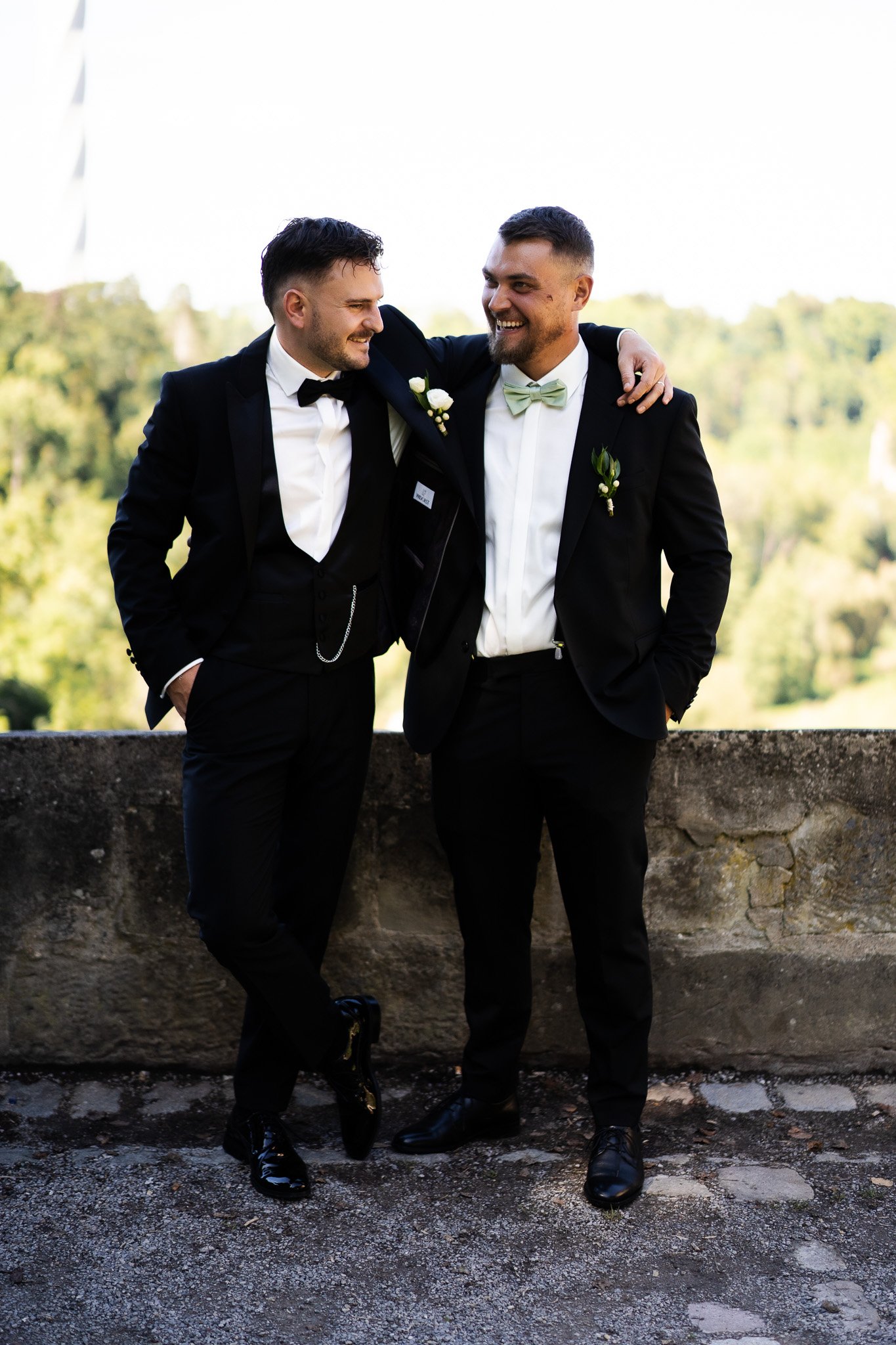 Zwei Männer in dunklen Anzügen feiern gemeinsam bei einem besonderen Anlass, vermutlich einer Hochzeit, vor einer natürlichen Kulisse mit Bäumen.