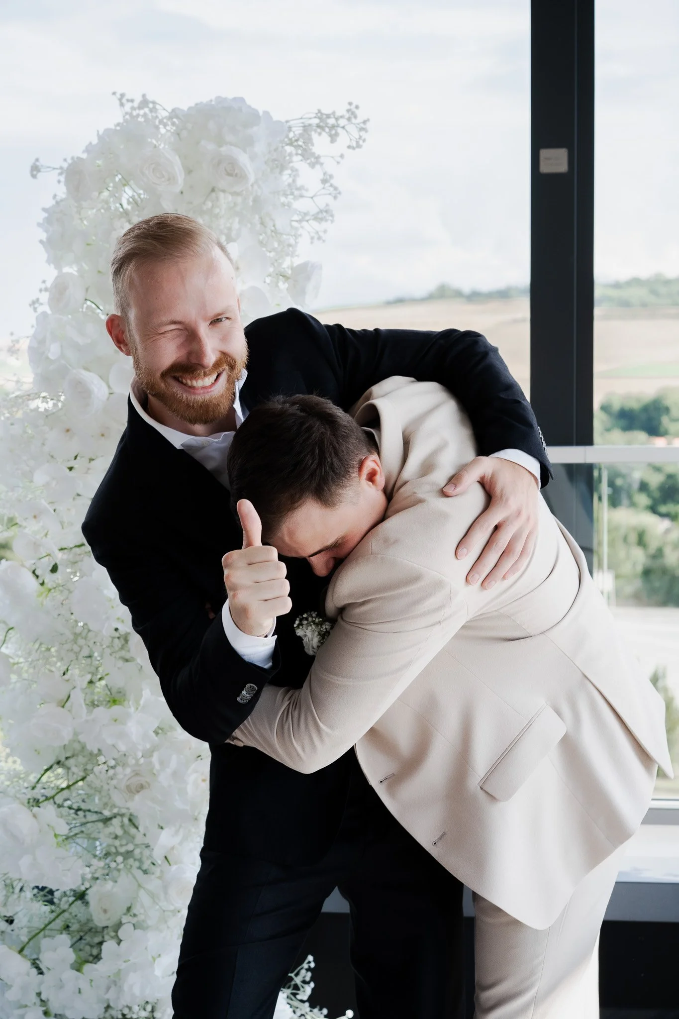 Zwei Männer in formeller Kleidung beim Umarmen, einer zeigt Daumen hoch, im Hintergrund weiße Blumen und eine große Fensterfront mit Blick nach draußen.