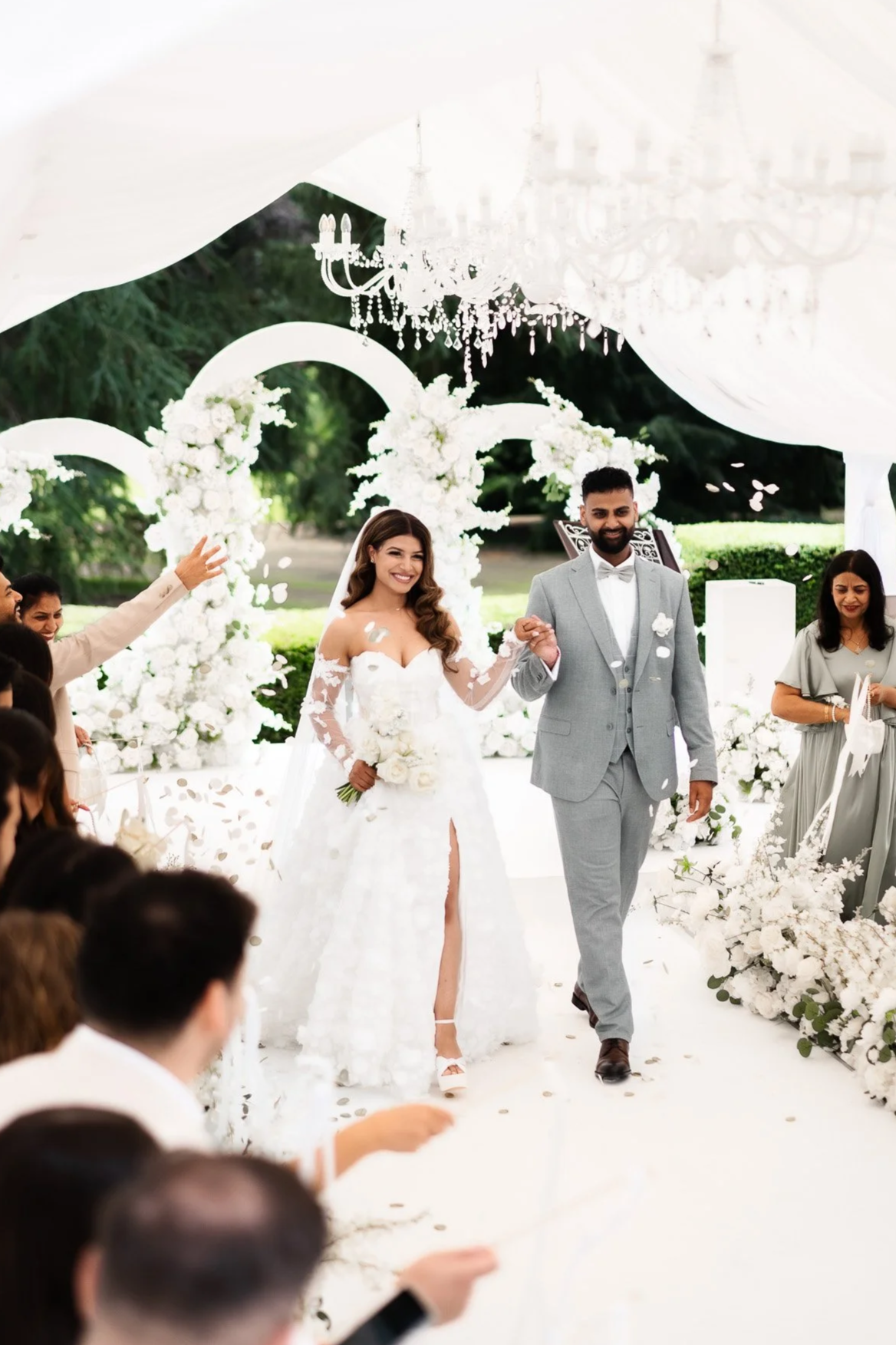 Ein Brautpaar bei ihrer Hochzeit, die Braut trägt ein weißes Kleid mit Schlitz, der Bräutigam trägt einen hellgrauen Anzug, sie gehen Hand in Hand unter einem weißen Hochzeitsthema mit Blumen und Kronleuchtern, Zuschauer feiern im Hintergrund.