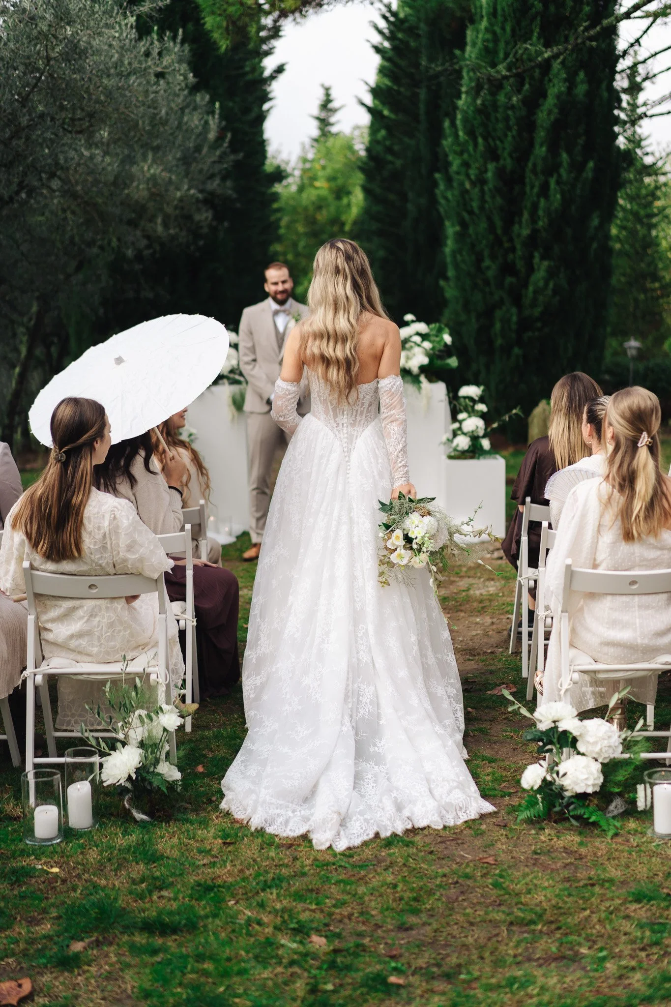 Eine Hochzeit im Freien mit einer Braut in einem weißen, spitzen Hochzeitkleid, die vor einem Altar steht. Der Bräutigam steht lächelnd in einem hellen Anzug. Gäste sitzen auf weißen Stühlen, einige halten Schirme und sehen zur Braut. Der Bereich ist