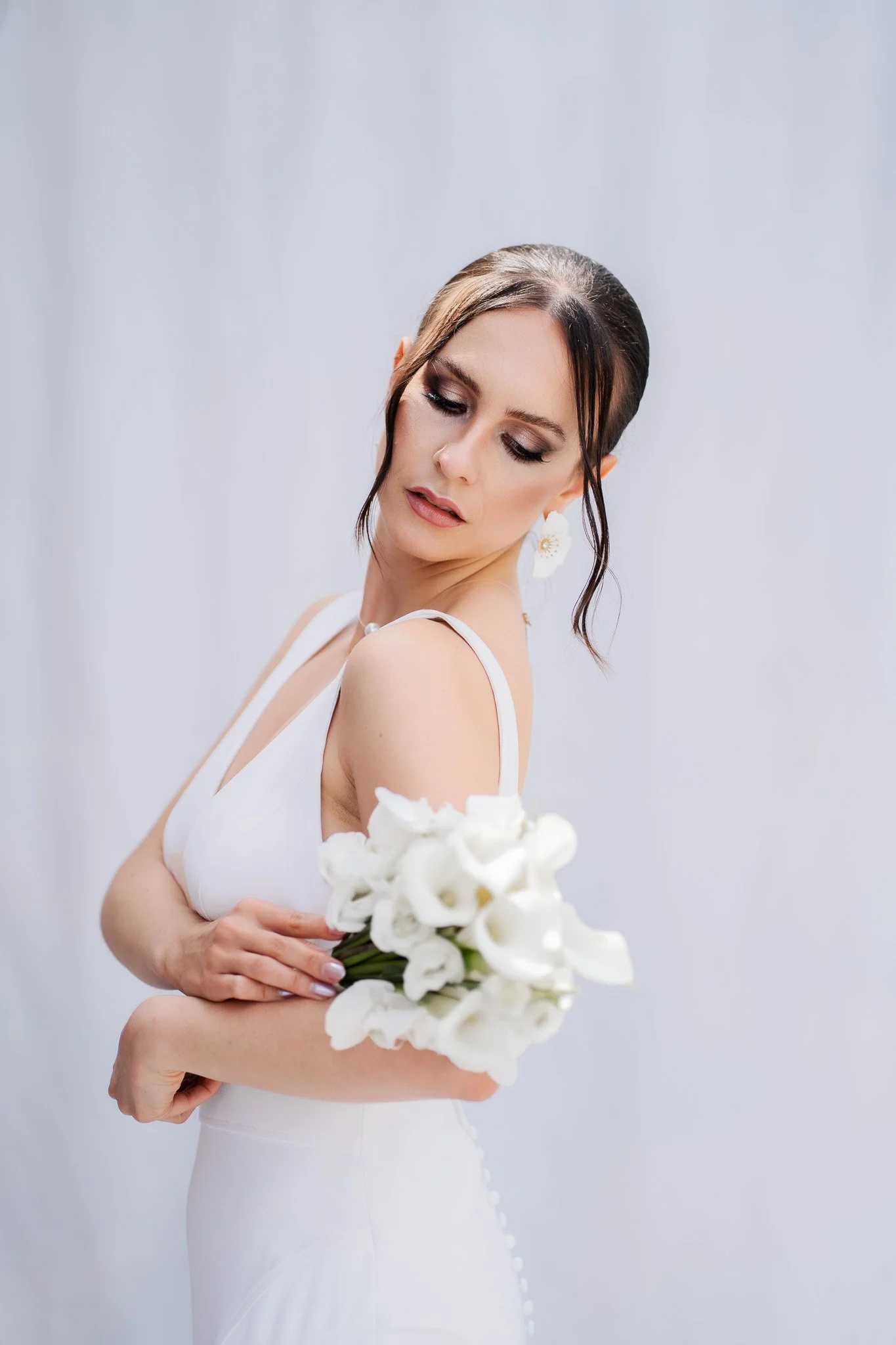 Junge Frau in Weißes Kleid hält weißen Blumenstrauß, Blick nach unten, festlich geschminkt, dunkle Haarupflege, Ohrschmuck, grauer Hintergrund.