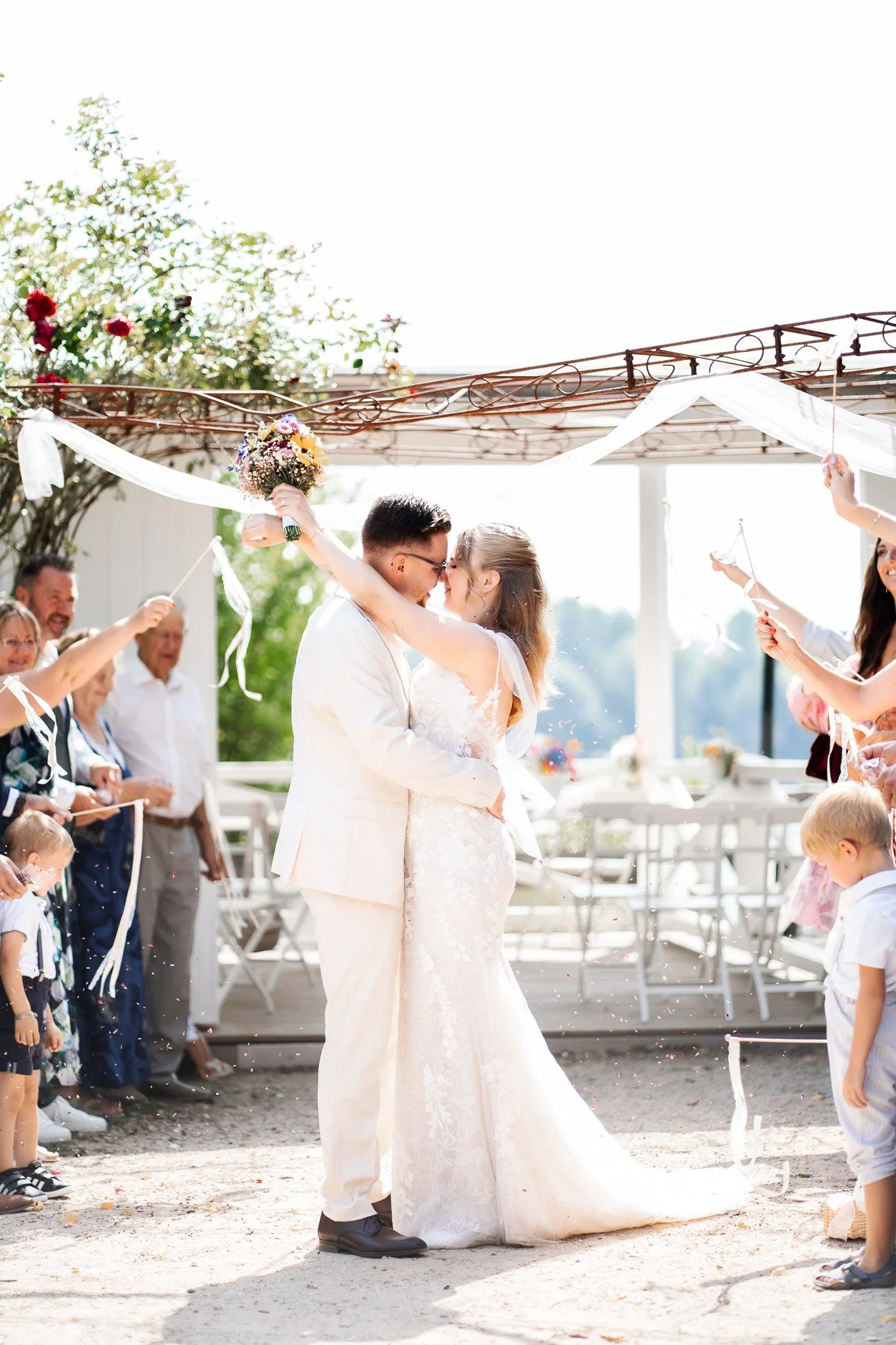 Das Brautpaar tanzt auf ihrer Hochzeitsfeier im Freien, umgeben von Freunden und Familie, bei Sonnenschein, während Konfetti geworfen wird.