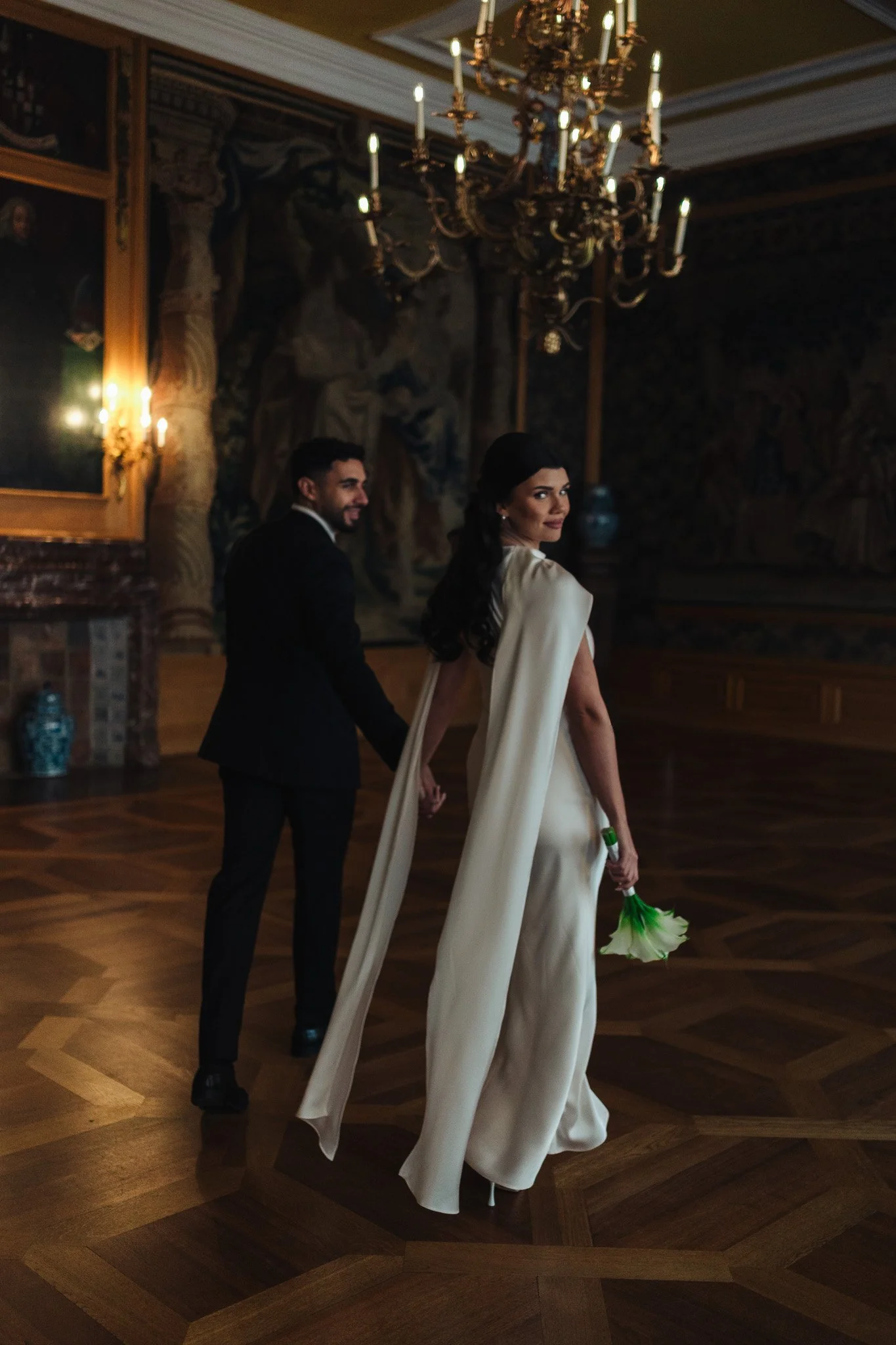 Ein Paar tanzt Hand in Hand in einem eleganten Saal mit Kronleuchter und Wandbildern, die den Raum schmücken. Die Frau trägt ein langes, cremefarbenes Kleid und hält einen weiß-grünen Blumenstrauß. Der Mann trägt einen schwarzen Anzug.