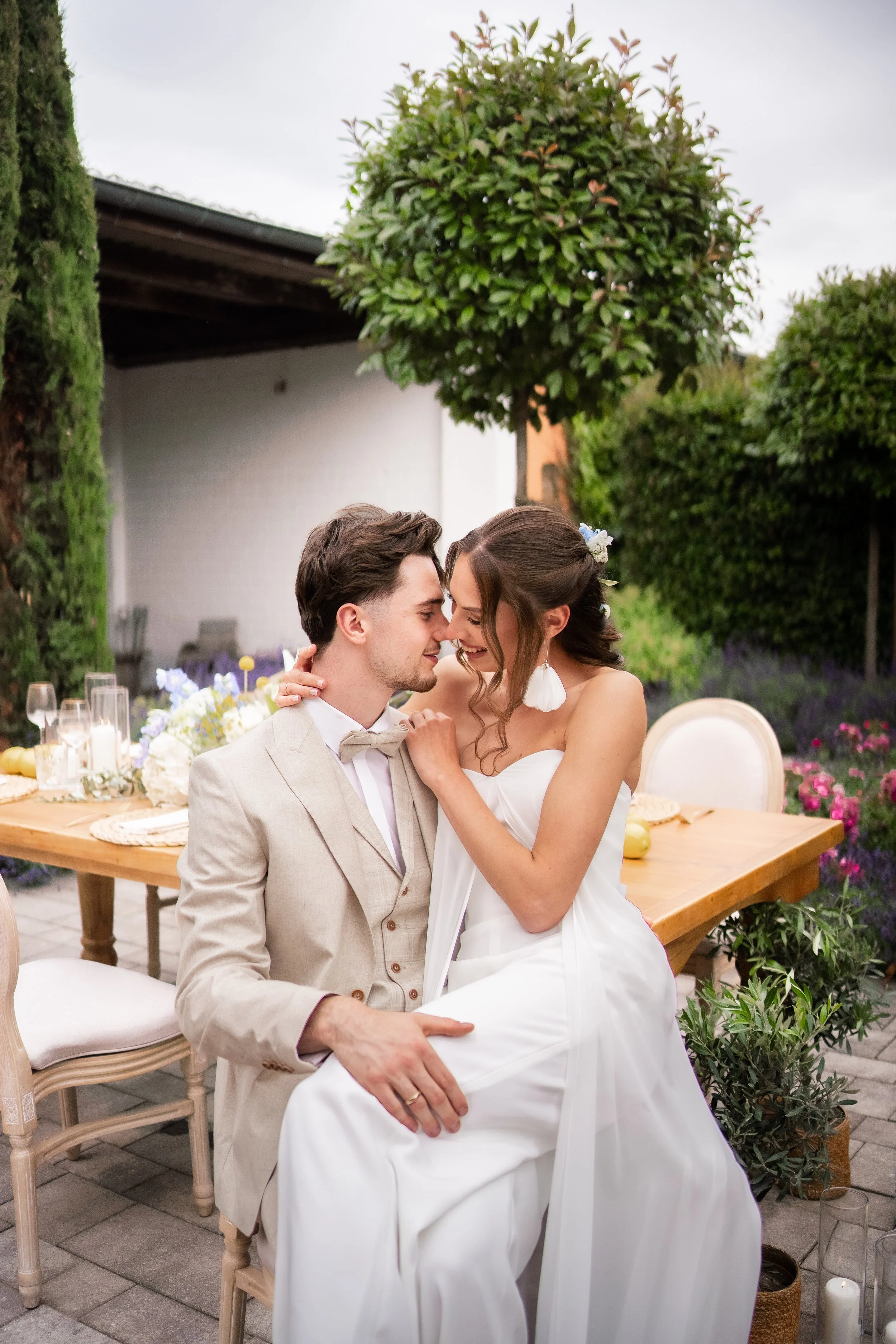 Ein Brautpaar in Hochzeitskleidung sitzt im Garten bei Abenddämmerung, umgeben von Blumen und Dekorationen, die Zweisamkeit und Liebe ausdrücken.