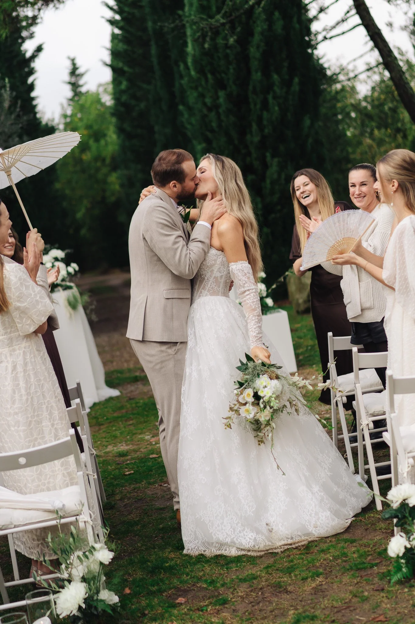 Ein heiratendes Paar beim Kuss, umgeben von lachenden Gästen bei einer Hochzeit im Freien.
