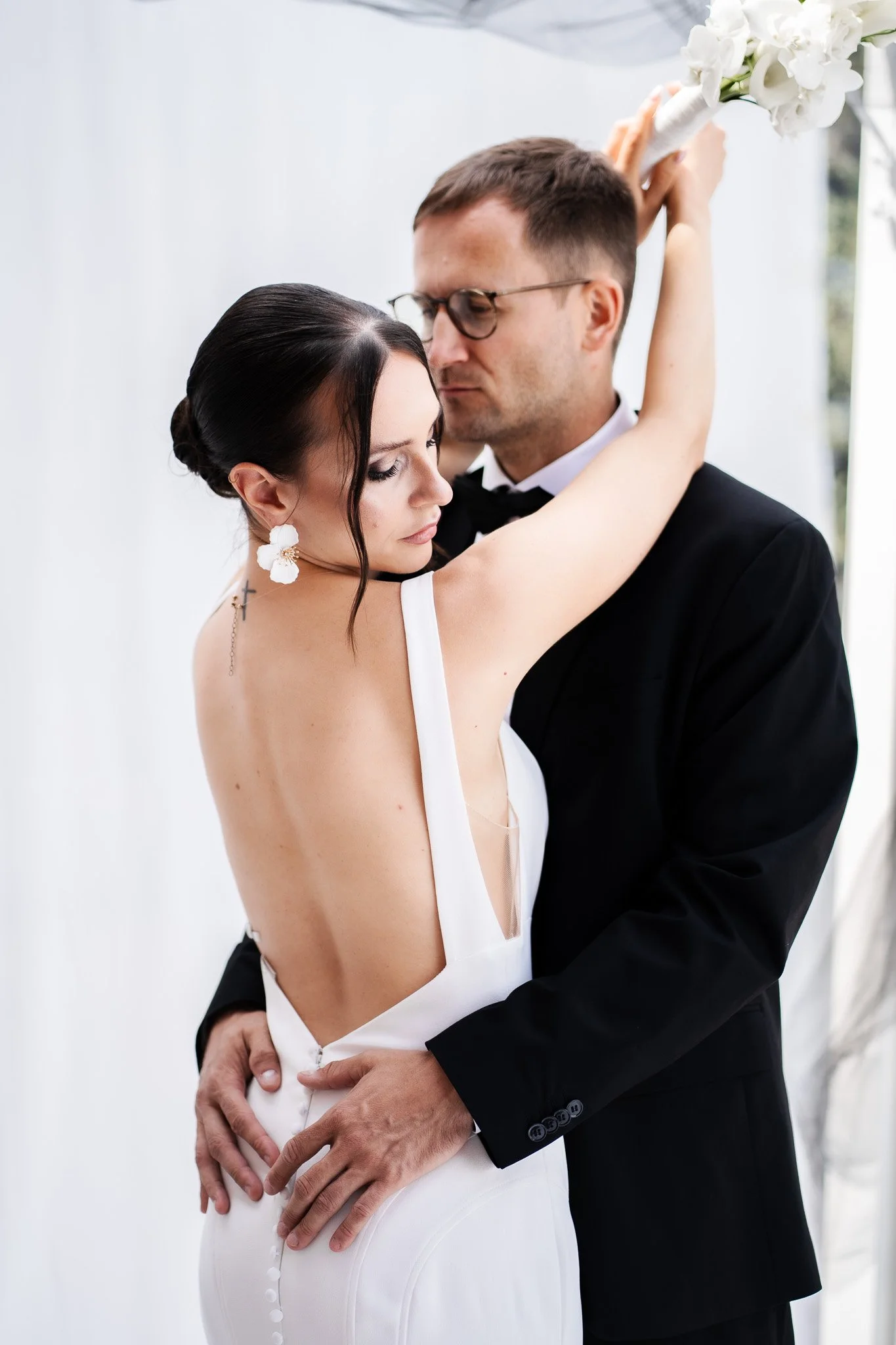 Ein Brautpaar bei einer Hochzeit, die Frau trägt ein weißes Kleid mit offenem Rücken und hält einen Blumenstrauß, während der Mann in einem schwarzen Anzug steht.