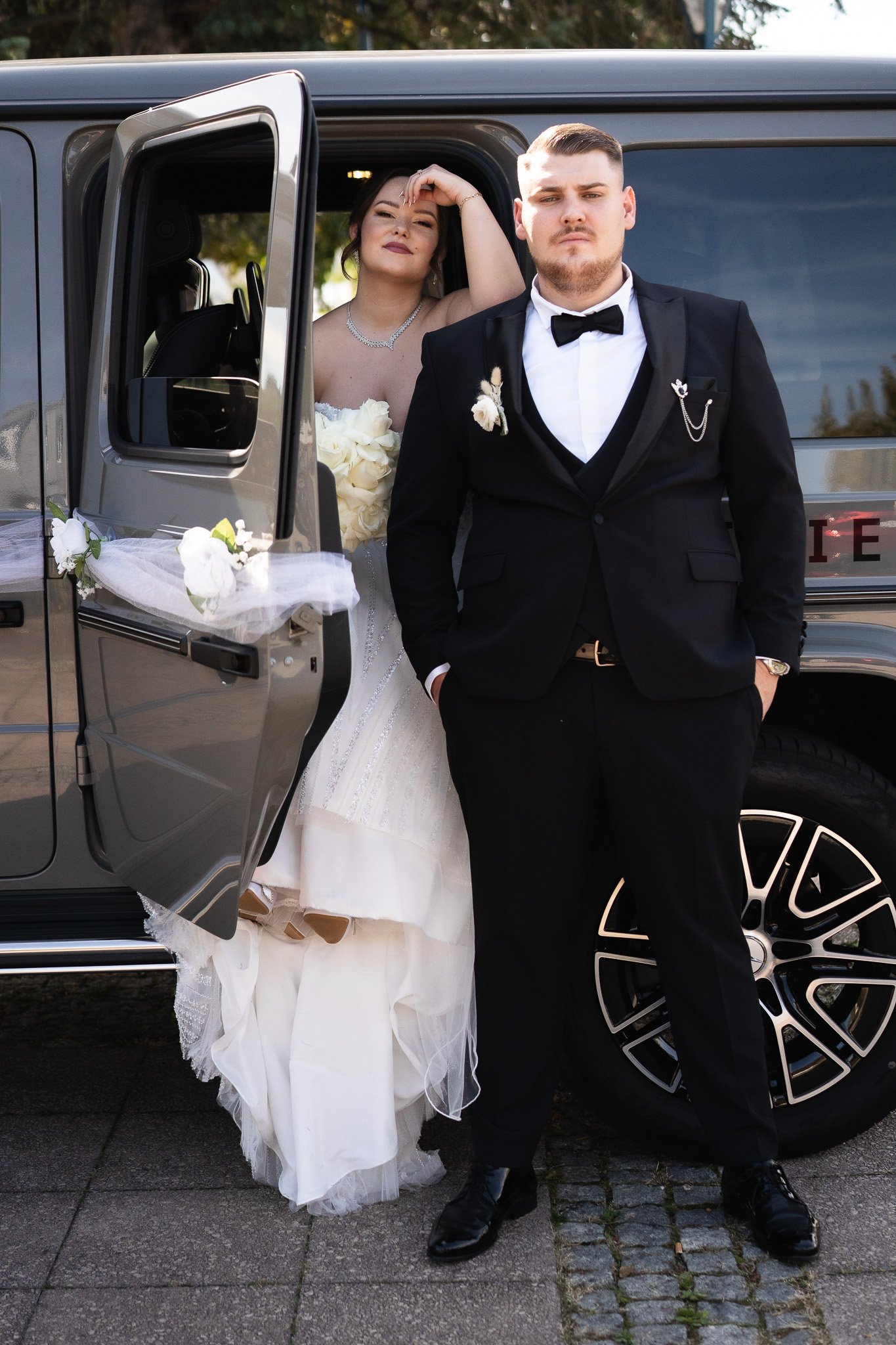 Ein Brautpaar steht vor einem schwarzen Fahrzeug, die Braut trägt ein weißes Hochzeitskleid und hält einen Blumenstrauß, der Bräutigam trägt einen schwarzen Anzug mit Fliege.