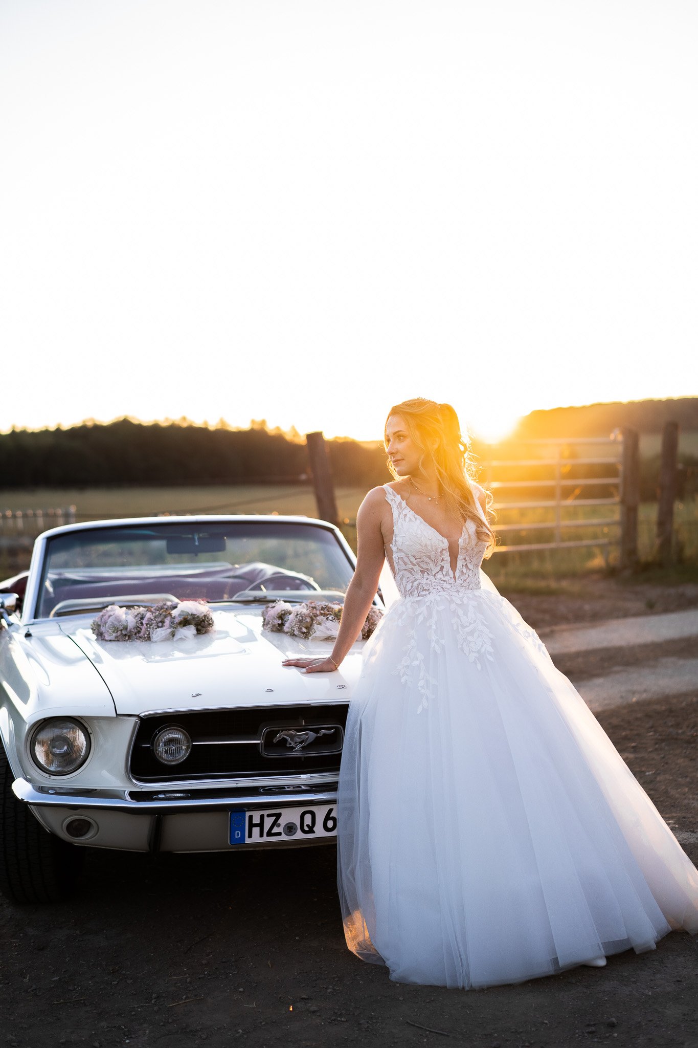 Braut in Hochzeitskleid neben einem weißen Ford Mustang Cabrio bei Sonnenuntergang auf dem Land.