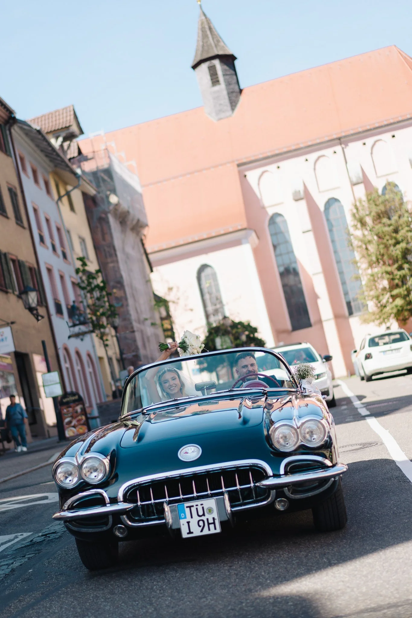 Ein junges Paar fährt in einem schwarzen Oldtimer-Cabrio durch eine belebte Stadtstraße, die Frau hält einen Blumenstrauß hoch. Im Hintergrund sind historische Gebäude mit farbigen Fassaden und einer Kirche zu sehen.