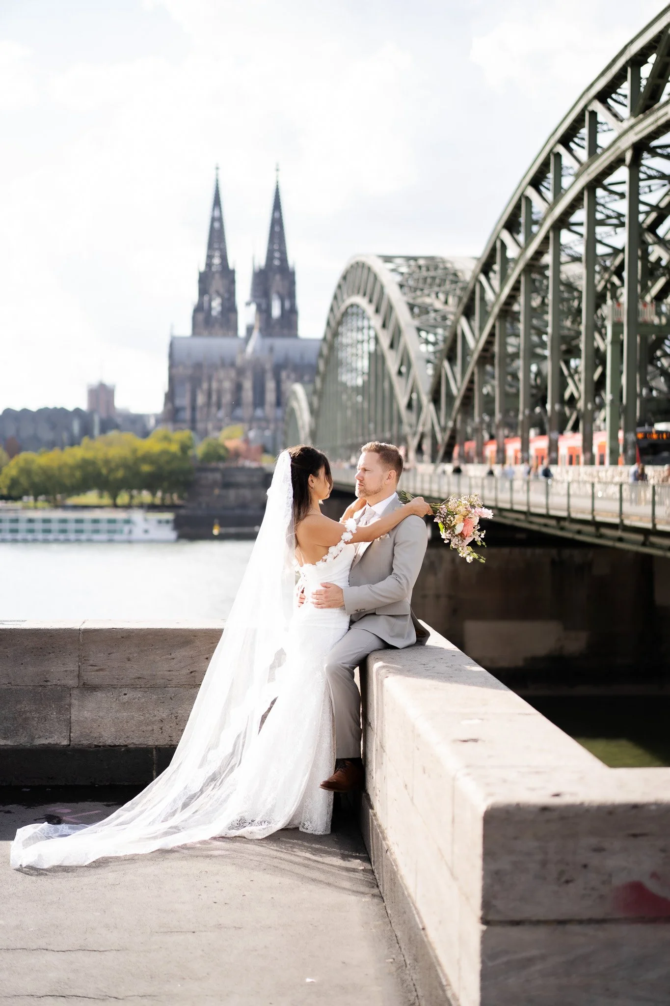 Ein Hochzeitsfoto eines Paares, das auf einer Mauer am Fluss in Köln sitzt, mit der Hohenzollernbrücke und dem Kölner Dom im Hintergrund bei Tageslicht