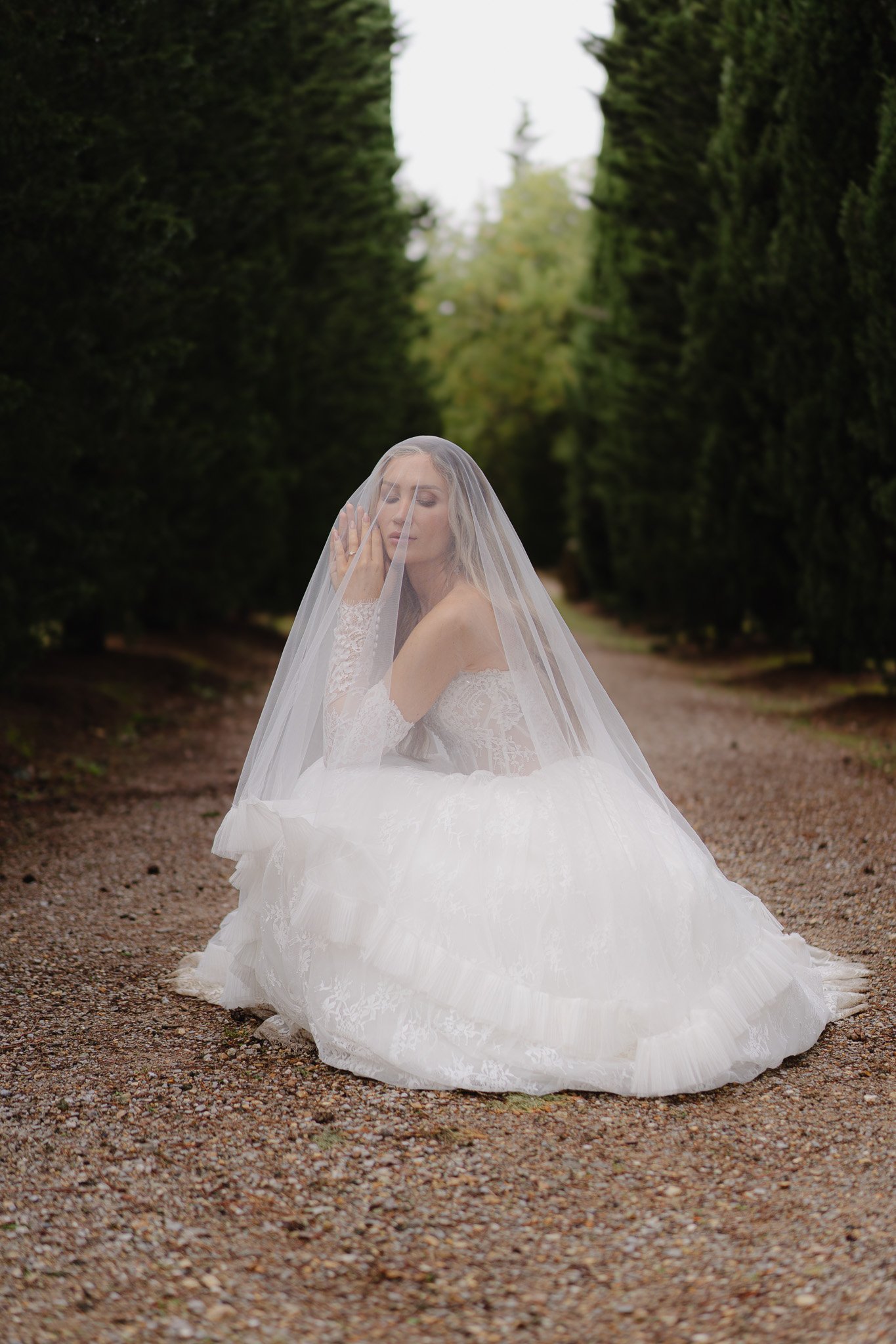 Junge Braut in Hochzeitskleid mit Schleier sitzt auf einem Kiesweg zwischen hohen, grünen Bäumen, umgeben von Natur.