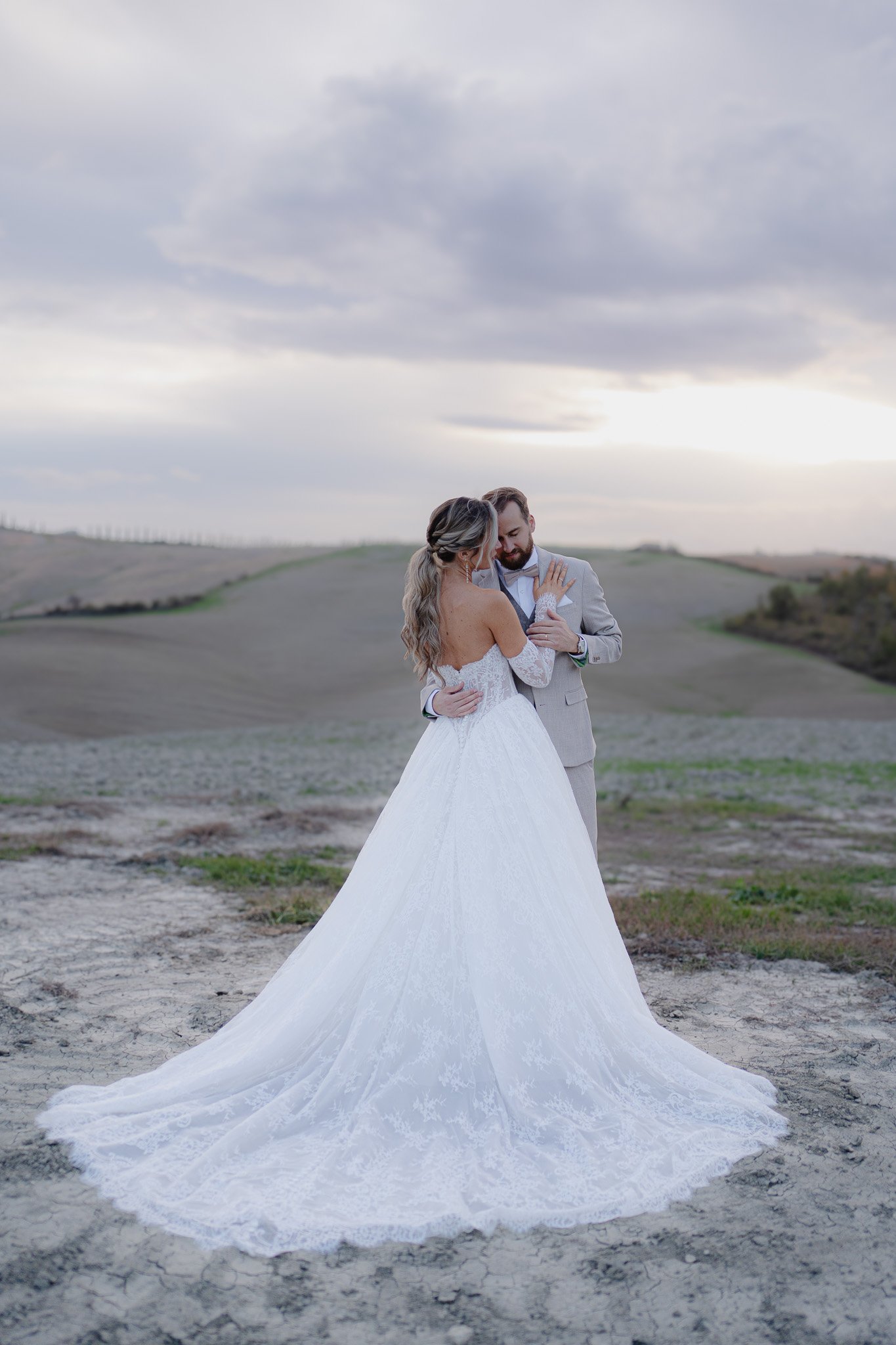 Ein Brautpaar in einer ländlichen, offenen Landschaft bei Sonnenuntergang, die Frau in einem weißen Brautkleid, der Mann in einem hellgrauen Anzug, umarmend und nahe beieinander stehend.