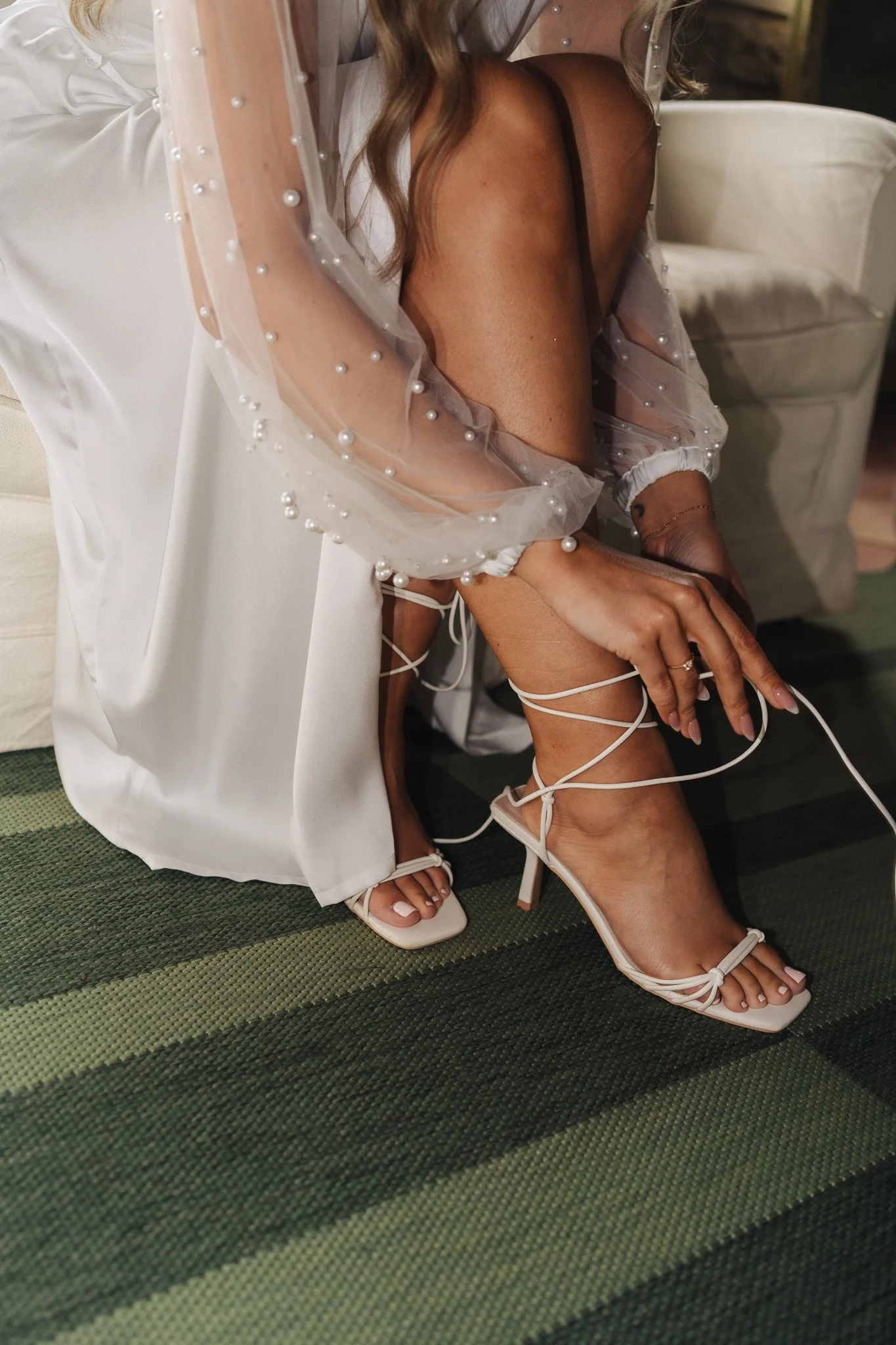 Nahaufnahme eines Frauenbeins in weißen hohen Schuhen mit Schnürung, eine andere Frau sitzt neben ihr, trägt ein schwarzes Kleid und eine transparente Jacke mit Perlen. Sie sitzt auf einem grünen Teppich. Im Hintergrund befindet sich ein weißes Möbel