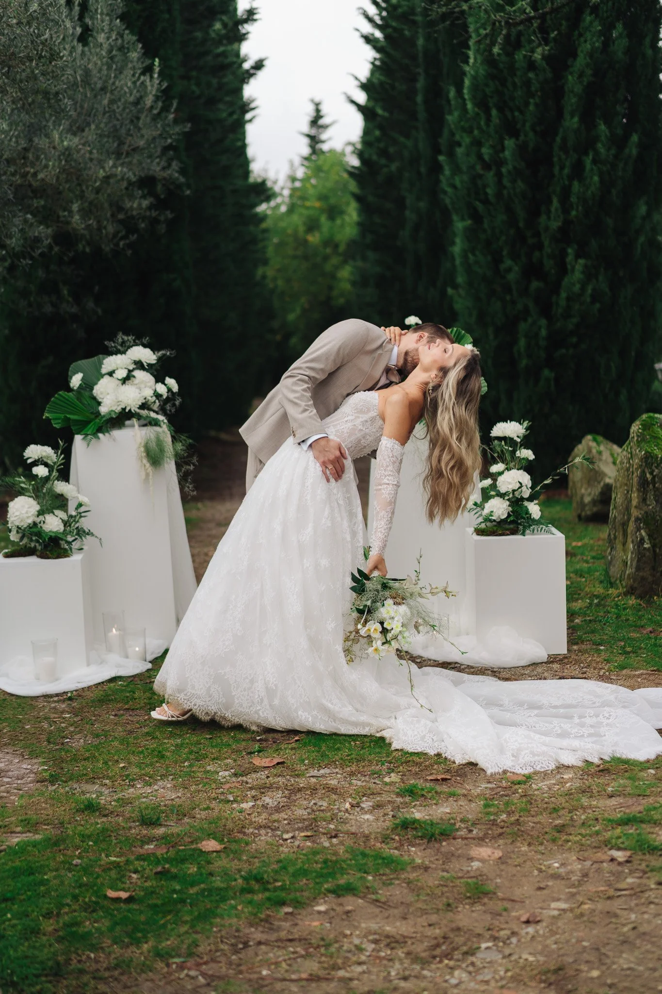 Ein Brautpaar tanzt oder küsst sich bei einer Hochzeit im Freien, umgeben von grünen Bäumen und weißen Dekorationen mit Blumen. Der Mann trägt einen hellen Anzug, die Frau ein weißes Brautkleid.