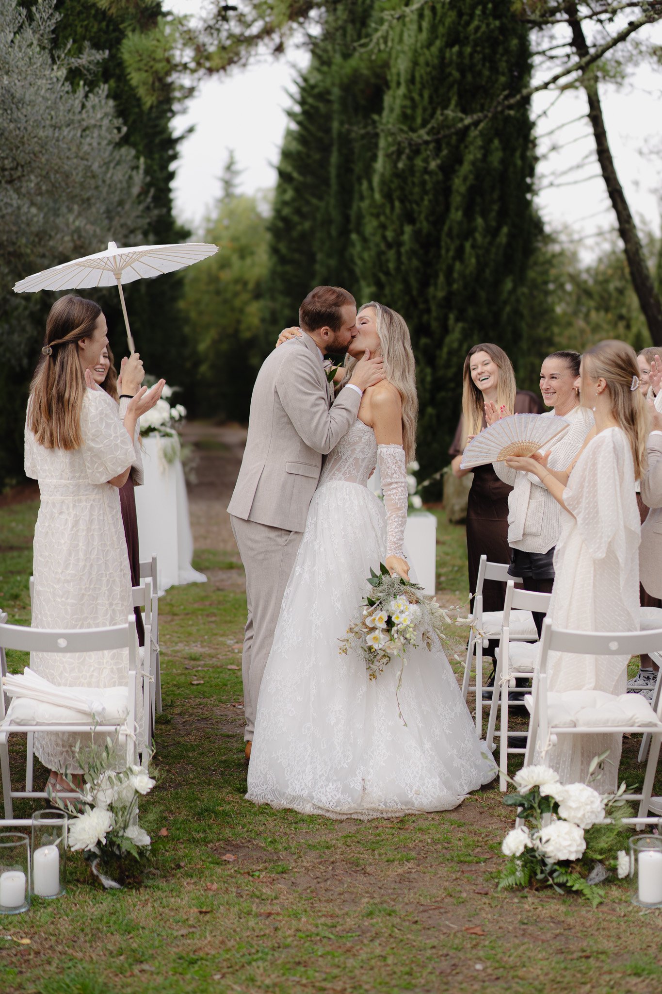 Ein Hochzeitsbild mit einem Paar, das sich küsst, während Gäste um sie herum lachen und applaudieren, im Freien in einer grün bewachsenen Umgebung.