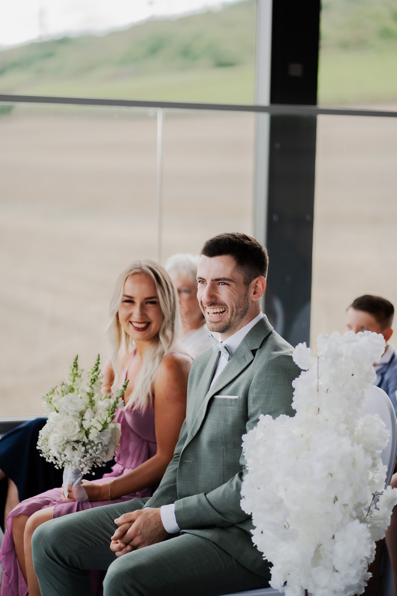 Ein Mann und eine Frau sitzen bei einer Hochzeitsfeier, beide lachen. Der Mann trägt einen grauen Anzug, die Frau hält einen Blumenstrauß. Im Hintergrund sind weitere Gäste und eine große Fensterfront, durch die eine ländliche Landschaft zu sehen ist