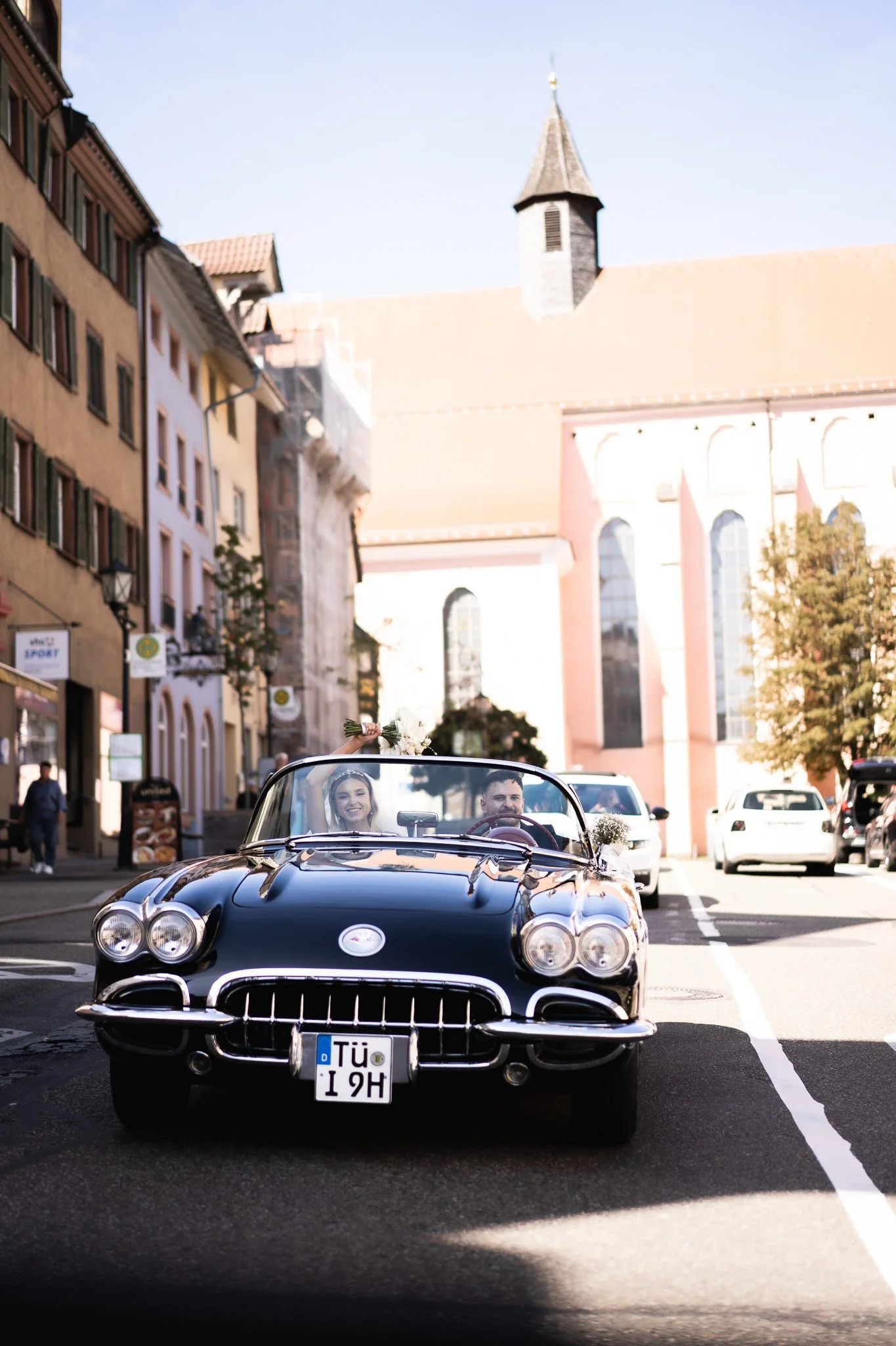 Ein älterer schwarzer Oldtimersportwagen fährt durch eine belebte europäische Stadtstraße, mit einem lachenden Paar im Innenraum, das wahrscheinlich Hochzeit feiert.