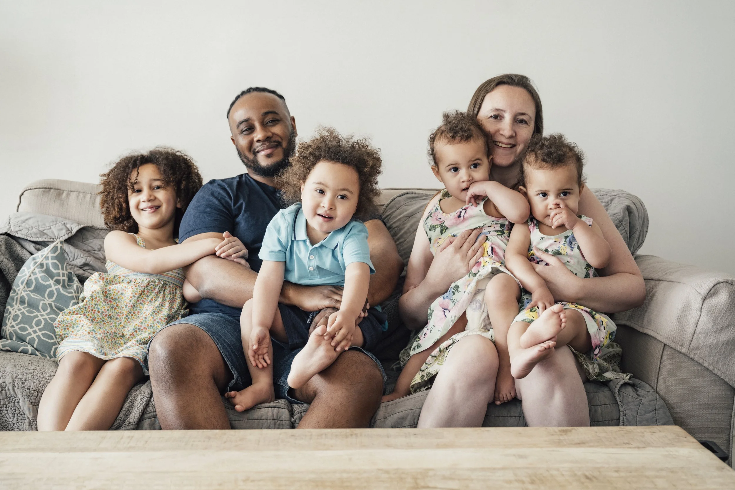 A happy multiracial family of four children, a mother, and a father sitting on a couch in a living room.
