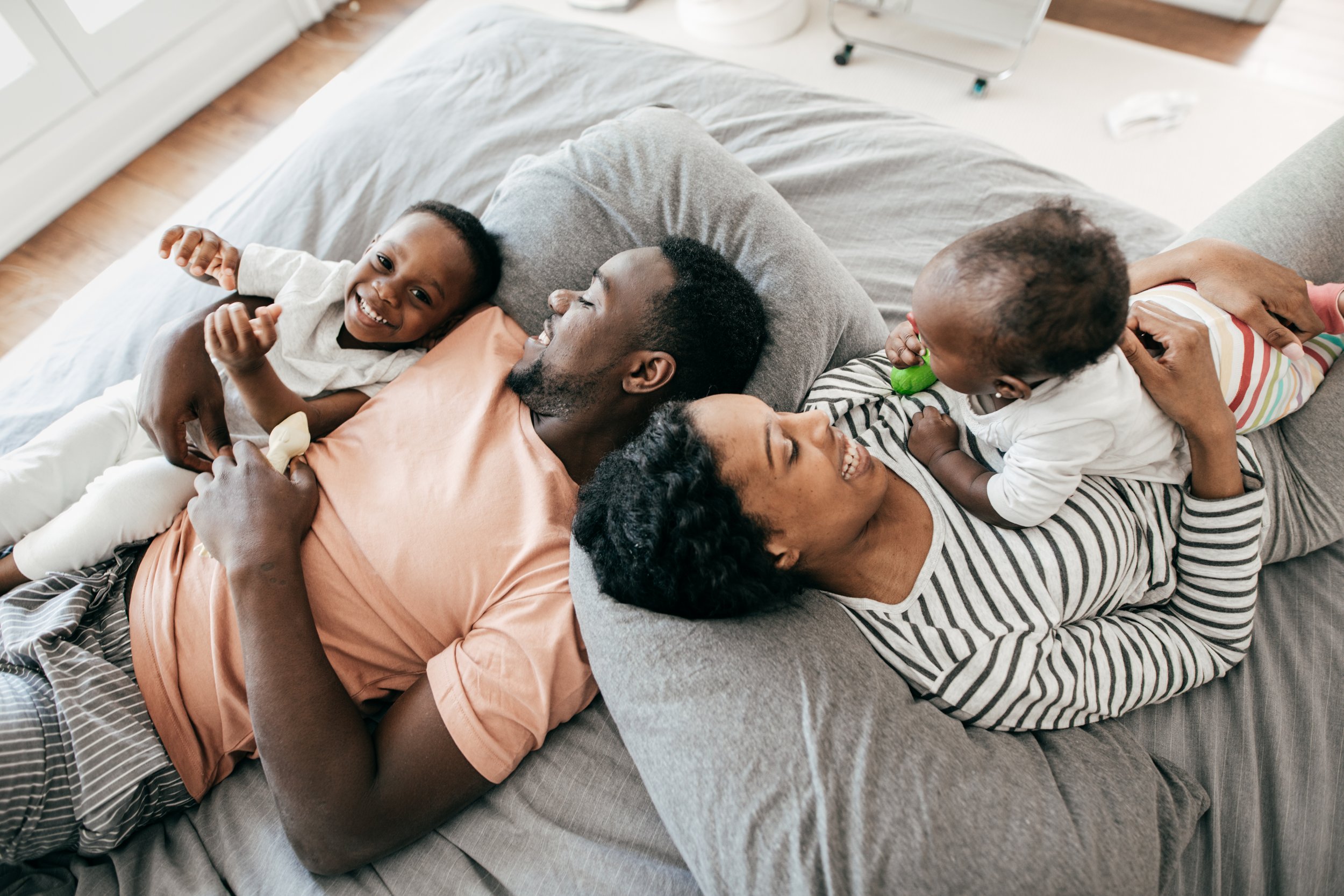 Family of four lying on bed, hugging and smiling, enjoying playful moment.