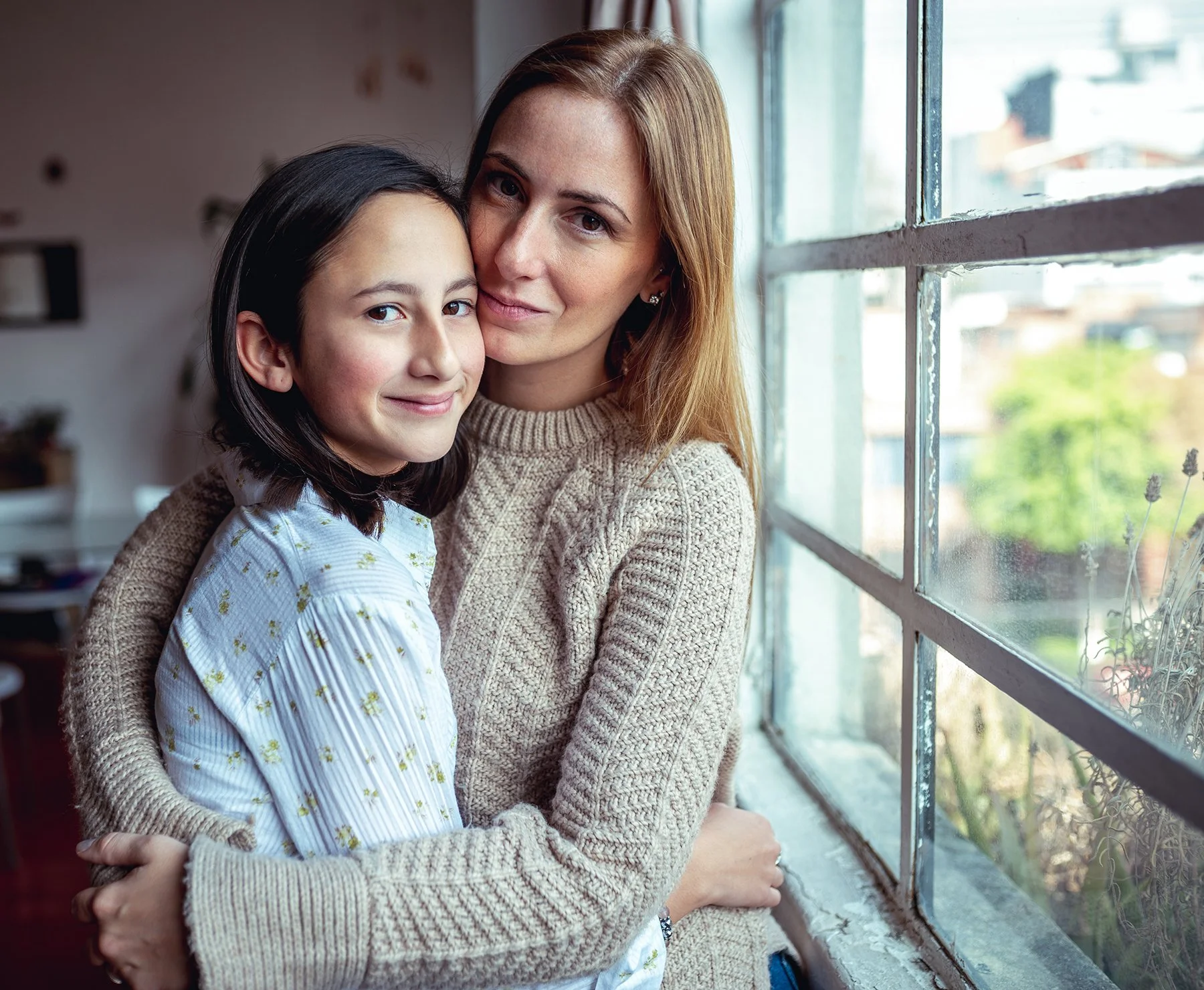 Mother and daughter hugging in front of a window.
