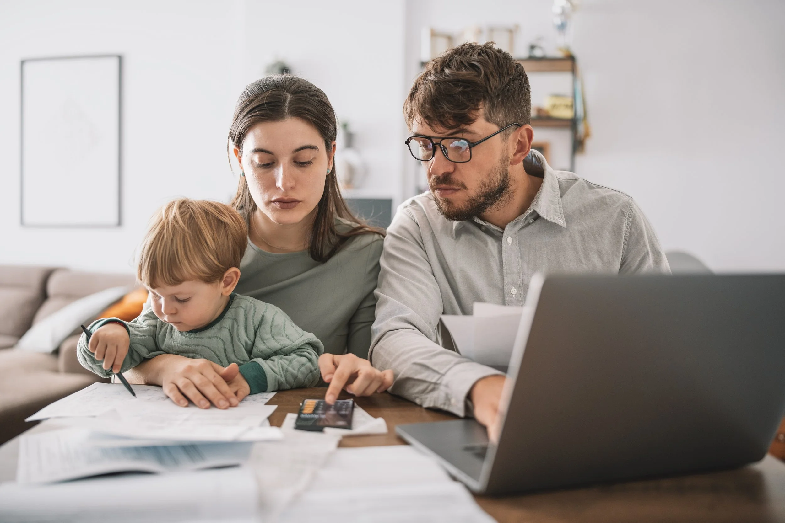 Young family of three with a toddler, sitting at a table managing their budget, paying bills and taxes online .