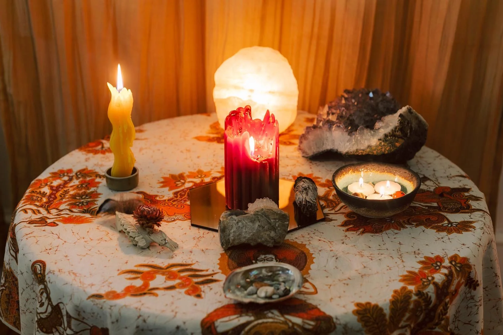 A round table with an orange, white, and red floral tablecloth. On the table, there are candles, rocks, and a salt lamp. There are four candles: a yellow one in a holder, a red one in the center, and three small white ones in a bowl. The background is a wood-paneled wall.