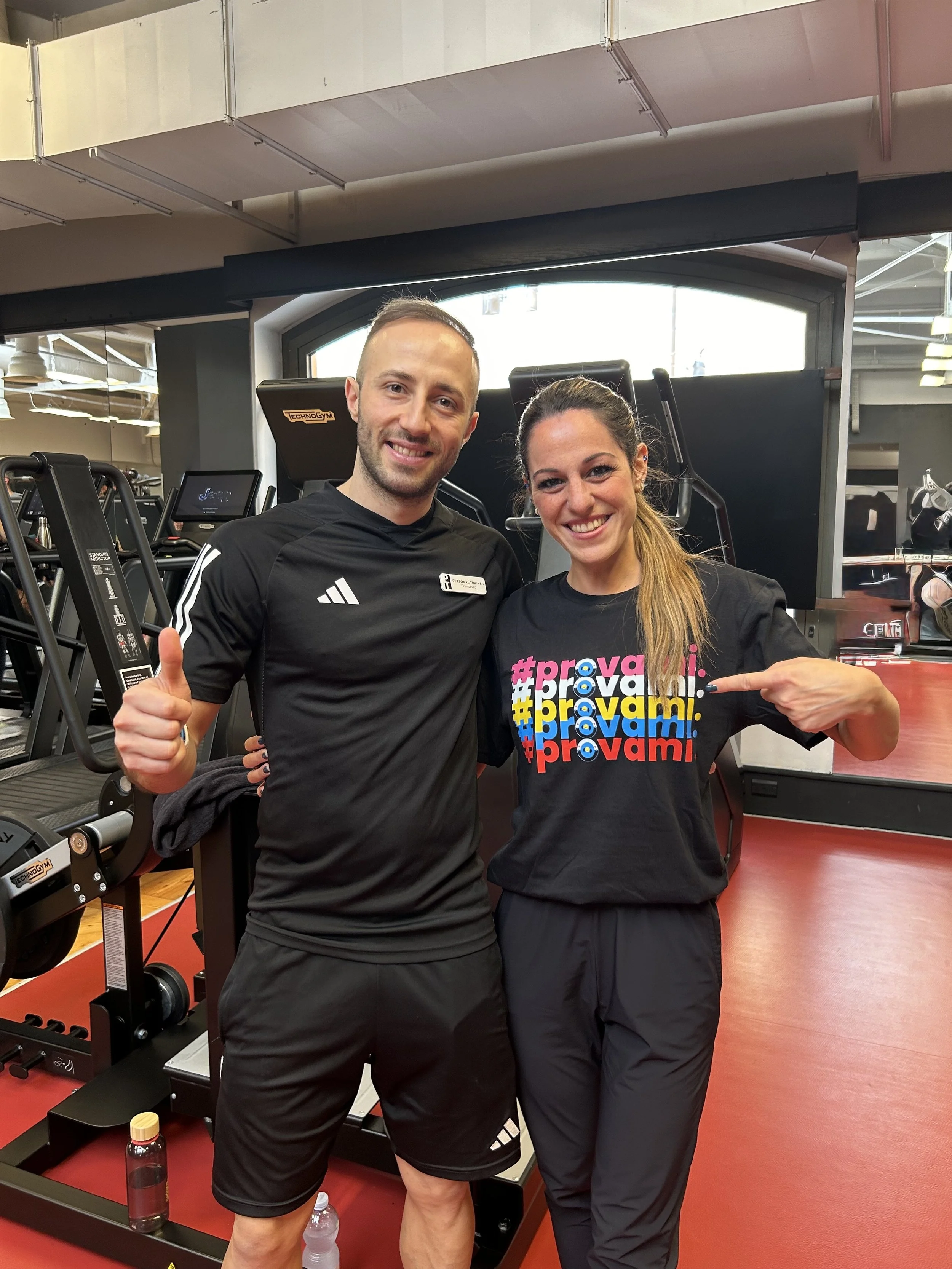Due persone sorridenti si trovano in una palestra, un uomo con maglietta nera e una donna con maglietta colorata con la scritta '#provami'.