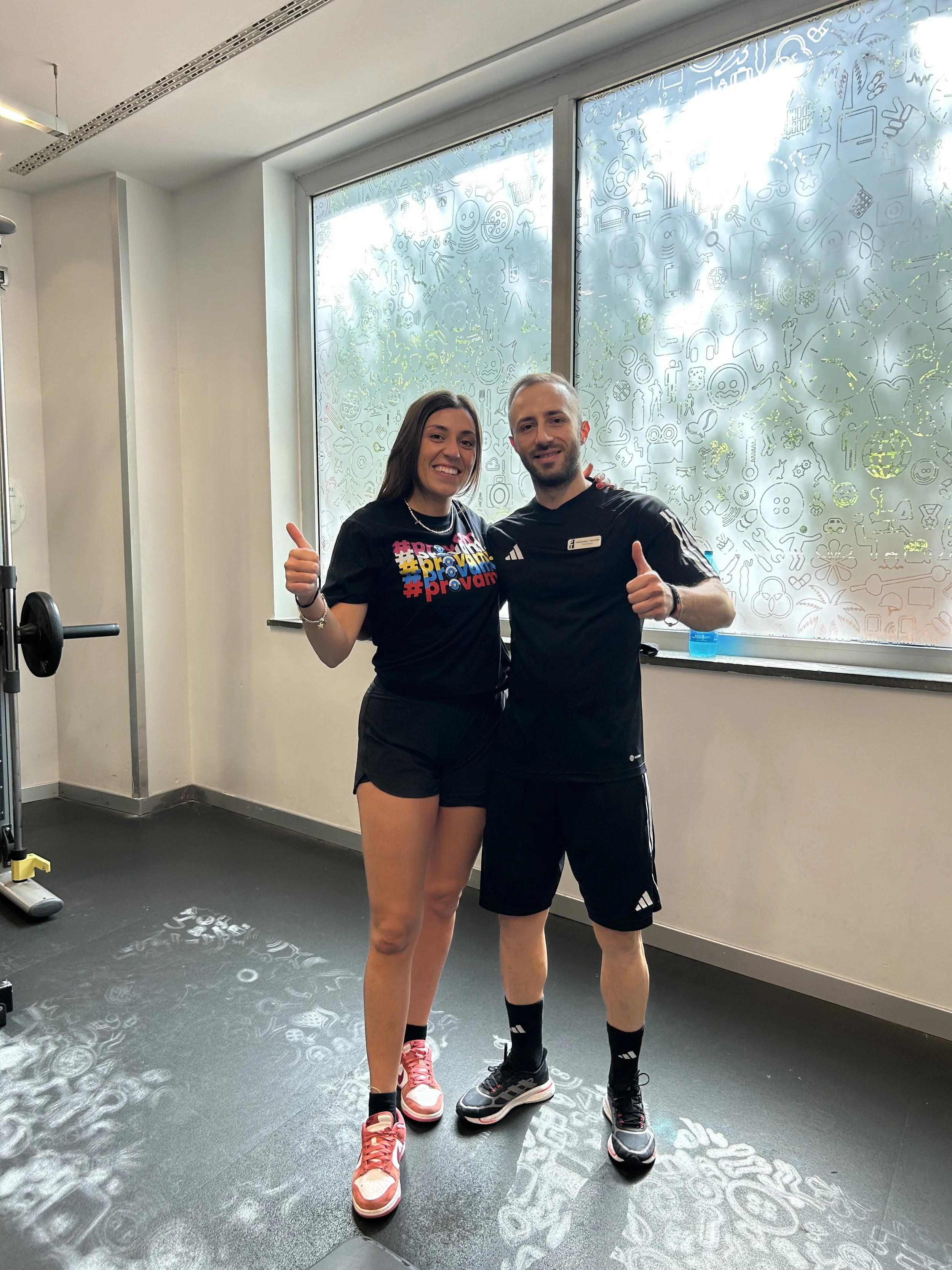 Due persone in abbigliamento sportivo sorridenti e che fanno il gesto del pollice in su, in una palestra.