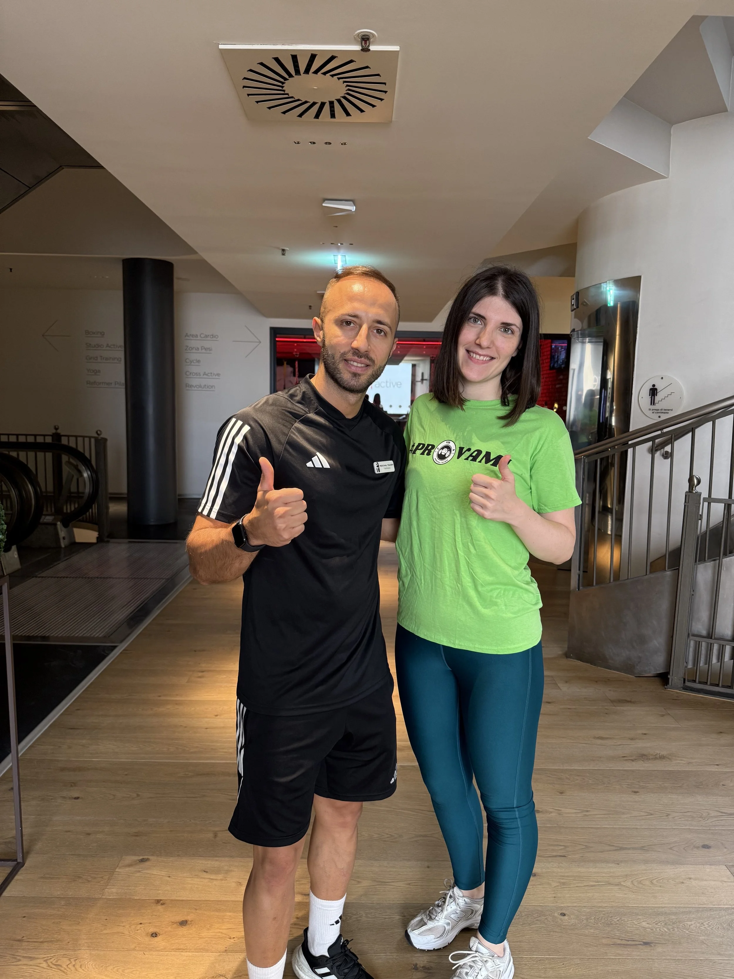 Due persone sorridenti e vestite da sport, un uomo con maglietta nera e una donna con maglietta verde, fanno un gesto di approvazione con il pollice in su, in un ambiente interno.