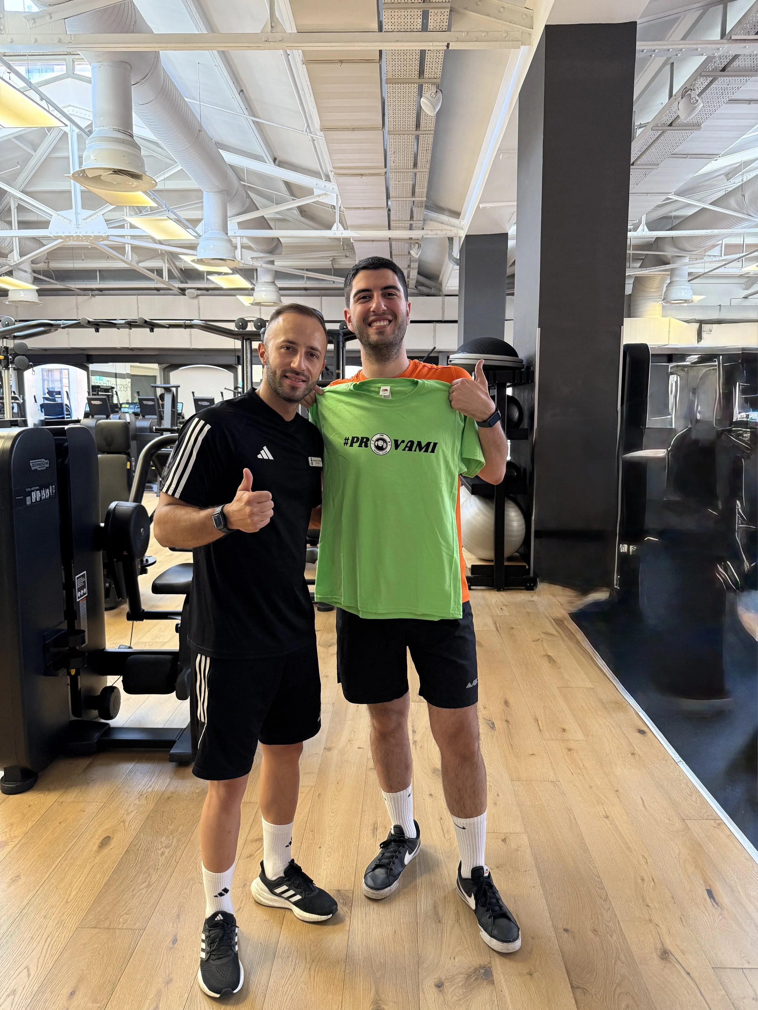 Due uomini in palestra, uno con maglietta nera e l'altro con una maglietta verde, sorridenti e che mostrano un pollice in su. Il secondo uomo tiene una maglietta verde con il testo '#PROVAMI'.