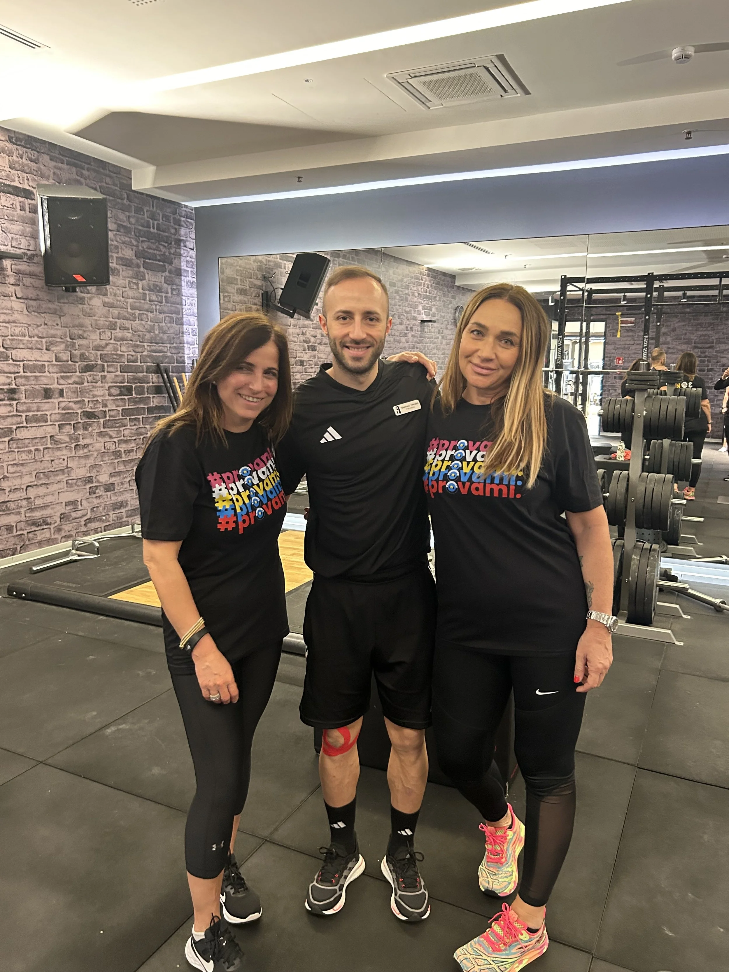 Tre persone sorridenti in palestra, due donne e un uomo, indossano abbigliamento sportivo e si abbracciano, con attrezzature per allenamento sullo sfondo.