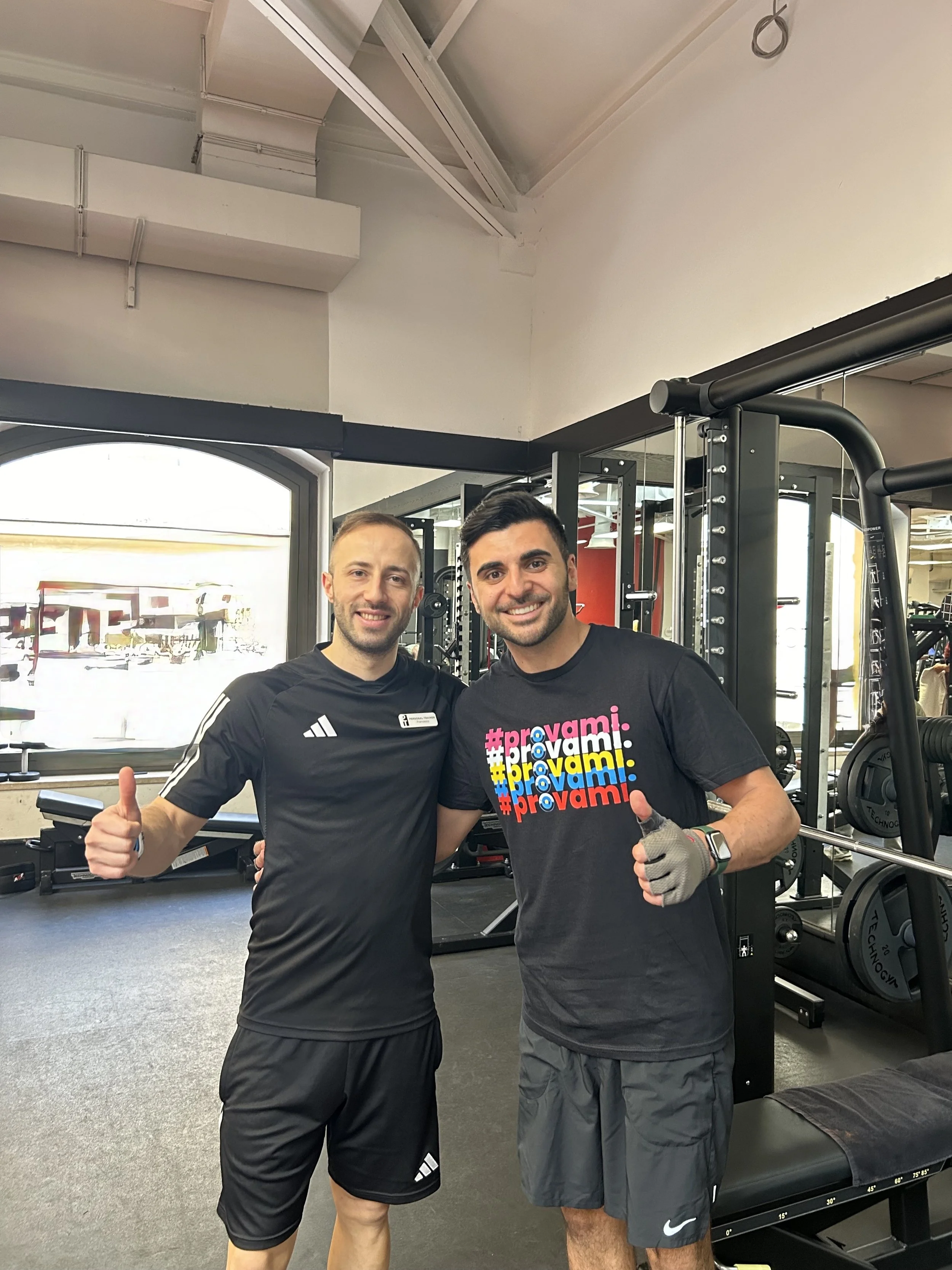 Due uomini sorridenti e sportivi, uno con una maglietta nera con logh Adidas e l'altro con una maglietta nera con scritto prova mi in colore rosa, grigio e blu, si trovano in palestra, posano con pollice alzato.
