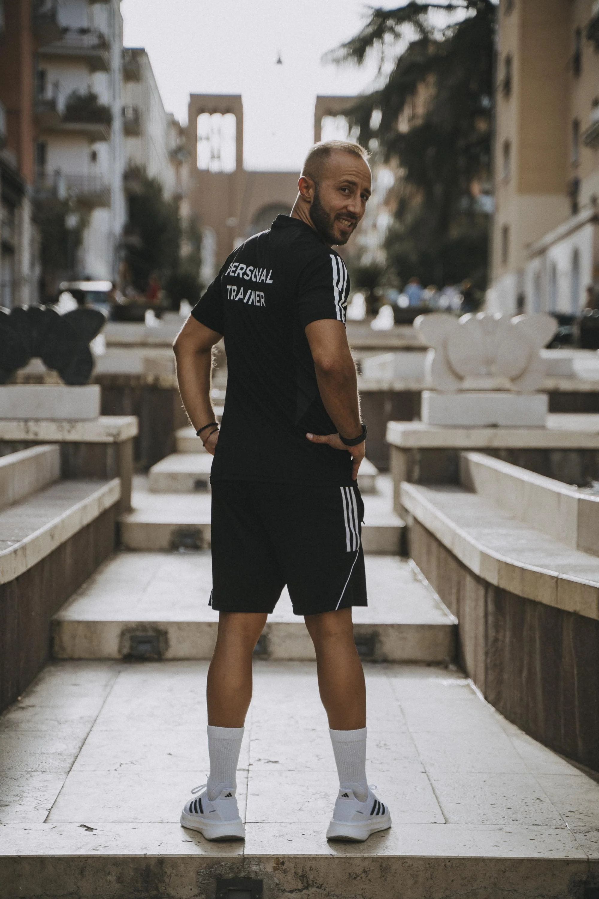Un uomo vestito in abbigliamento sportivo si trova davanti a una fontana in una zona urbana durante il tramonto, con edifici e un arco sullo sfondo.