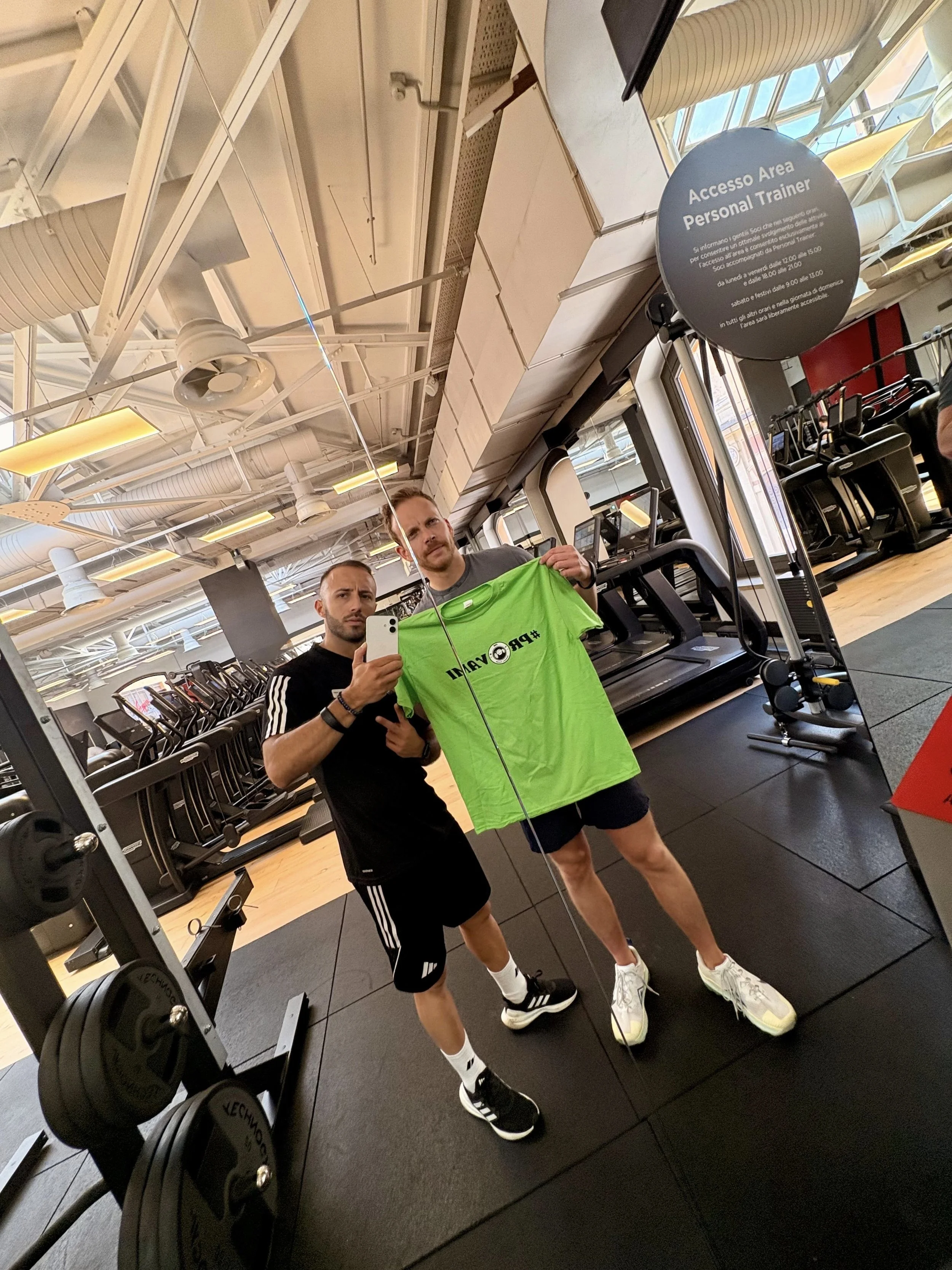 Due uomini si riflettono in uno specchio di una palestra, uno di loro tiene in mano una maglietta verde e si scatta un selfie in palestra.