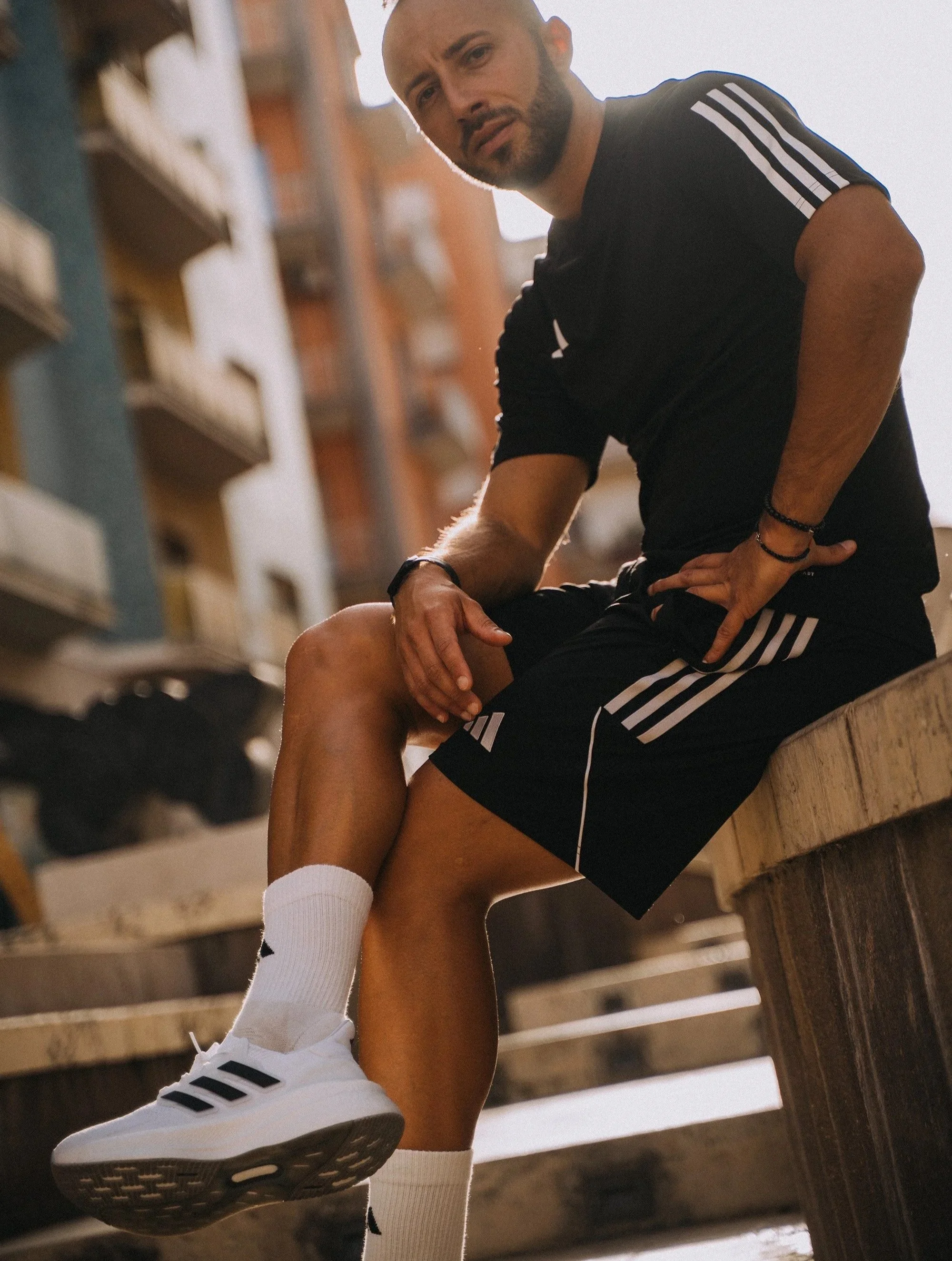 Uomo seduto su una panca di legno, si tiene un ginocchio con la mano e sembra indossare abbigliamento sportivo, incluso scarpe da ginnastica adidas e calze bianche.