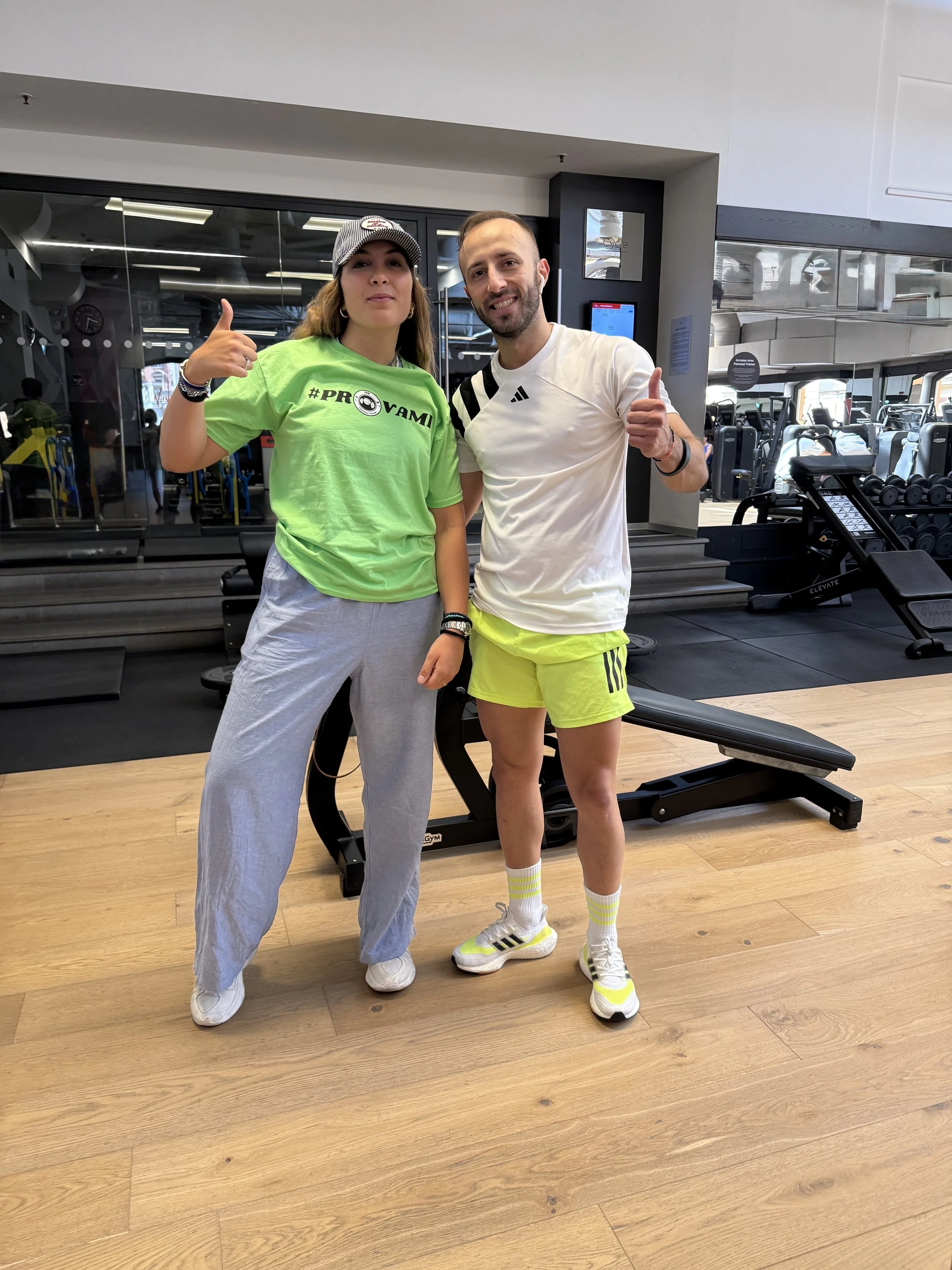 Due persone, una donna con abbigliamento sportivo verde e pantaloni comodi, e un uomo con maglietta bianca e pantaloncini corti gialli, stanno insieme in palestra, sorridendo e facendo il gesto del pollice in su.
