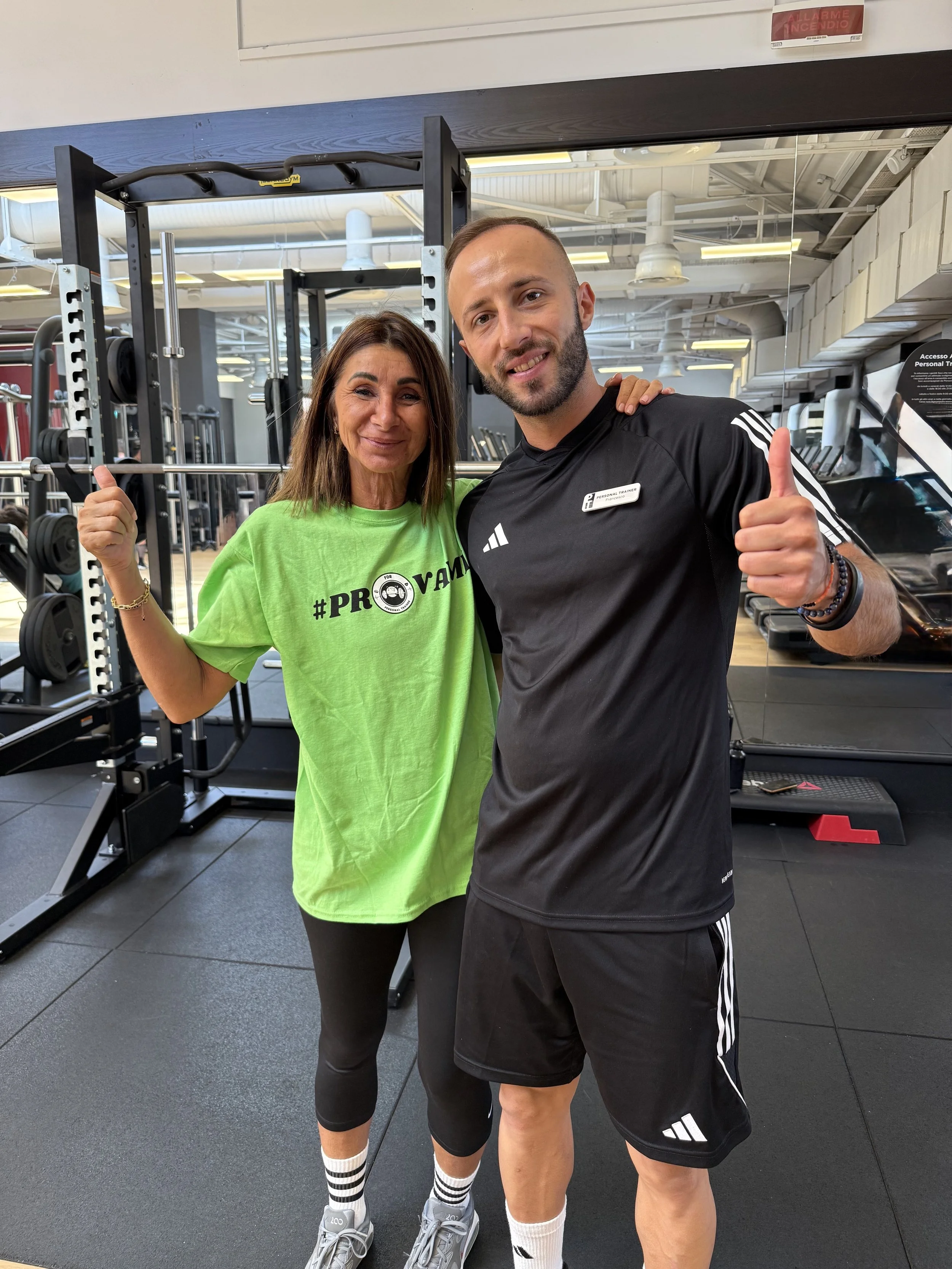 Una donna e un uomo si trovano in una palestra, sorridenti e che fanno il gesto del pollice in su. La donna indossa una maglietta verde con hashtag #PROVAM, pantaloni neri e scarpe sportive. L'uomo indossa una maglietta nera con logo e pantaloncini neri. Sullo sfondo ci sono attrezzi da palestra.